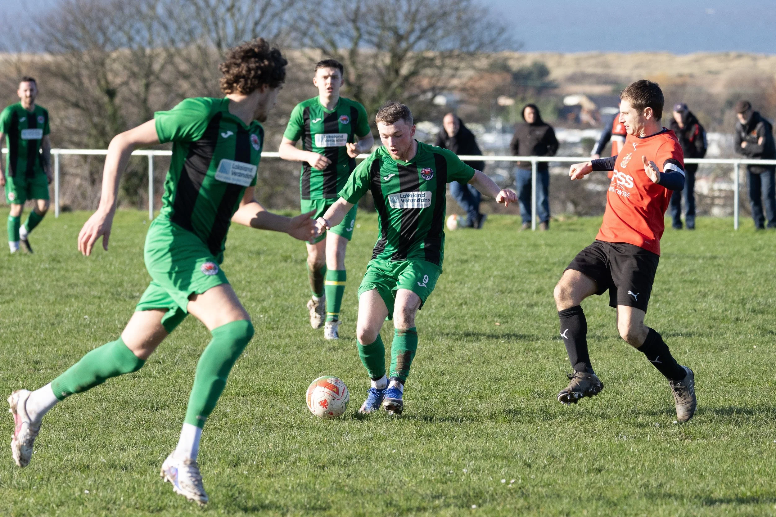Soccer players in green and black uniforms competing for the ball against a player in red and black uniform on a grassy field with a few spectators in the background.