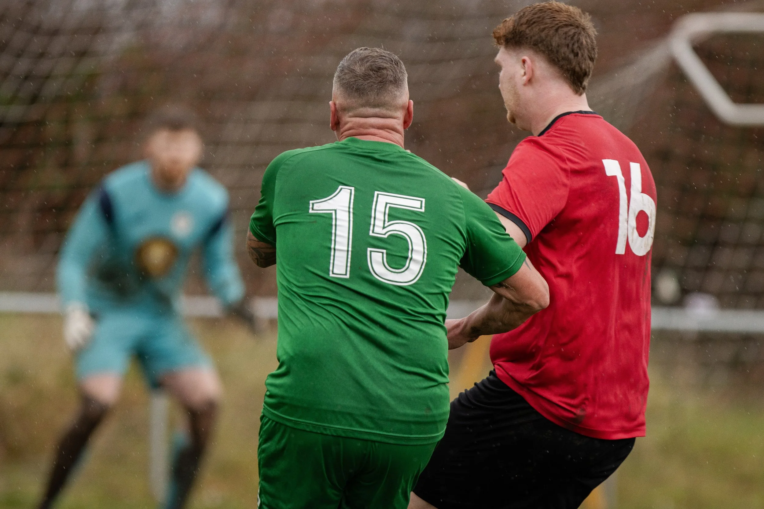 Two soccer players in a physical altercation on the field, with a goalkeeper in the background and a soccer goal net behind them.