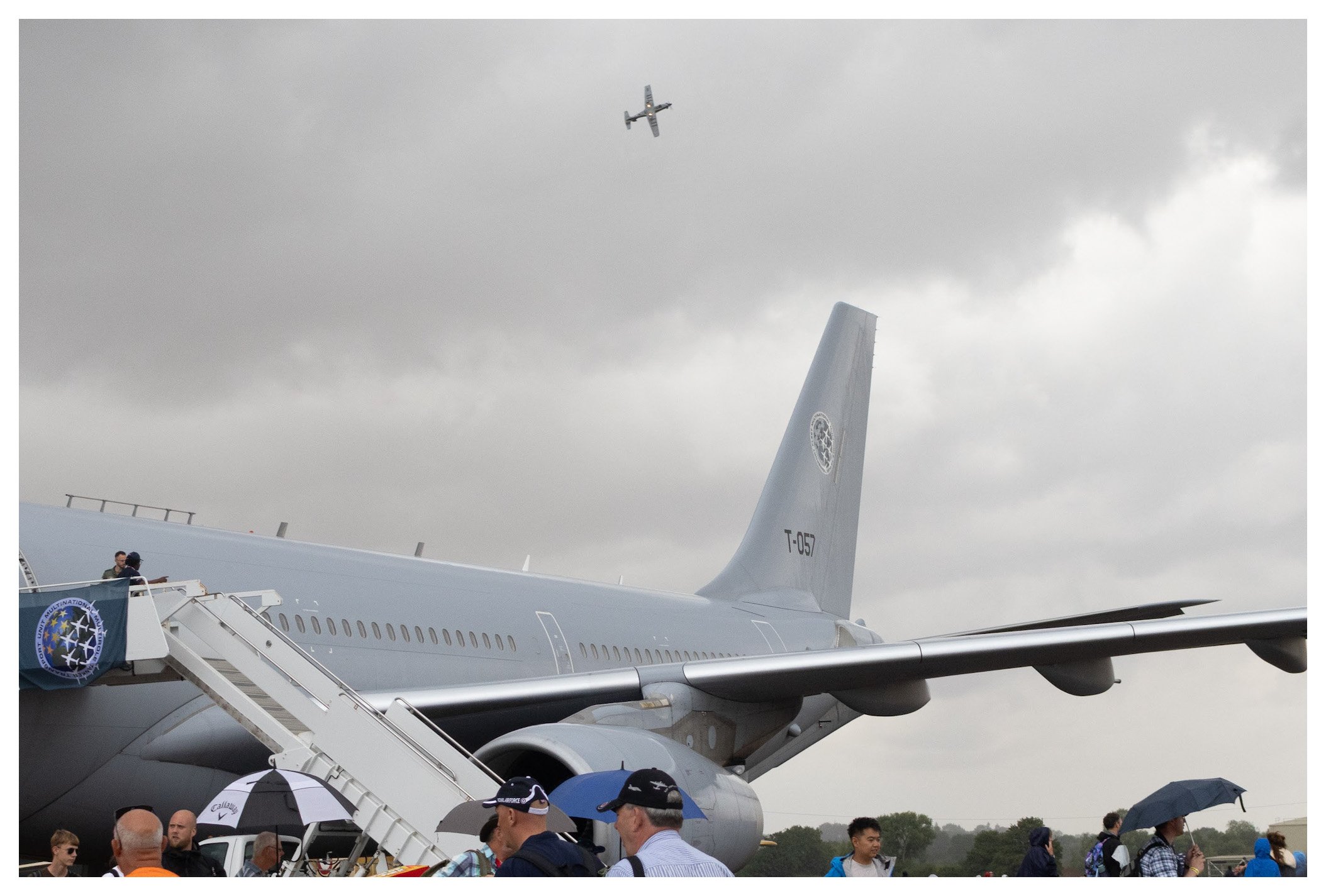 People boarding and walking around a large gray airplane with the tail number T-057, on an overcast day. An aircraft is flying overhead against a cloudy sky.