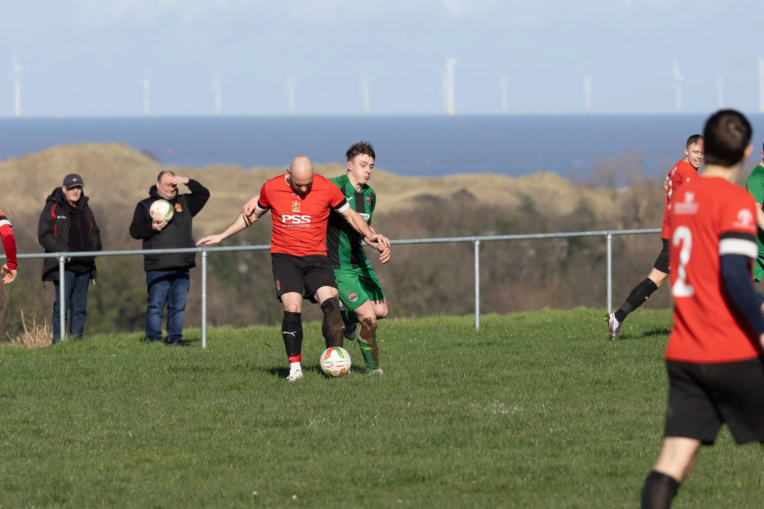 Two soccer players, one in a red jersey and one in a green jersey, competing for the ball on a grassy field during a match, with spectators in the background and wind turbines in the distance.