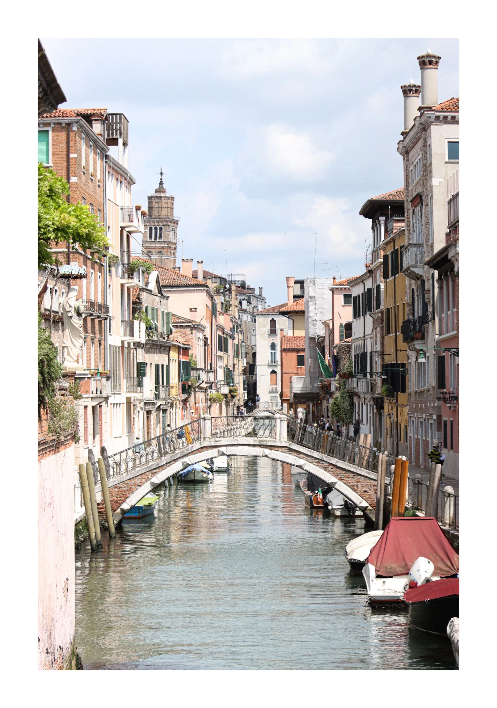 A canal with boats and a small stone bridge in a city with narrow buildings, some with balconies, on either side of the canal. There is a tall tower in the background and the sky is partly cloudy.