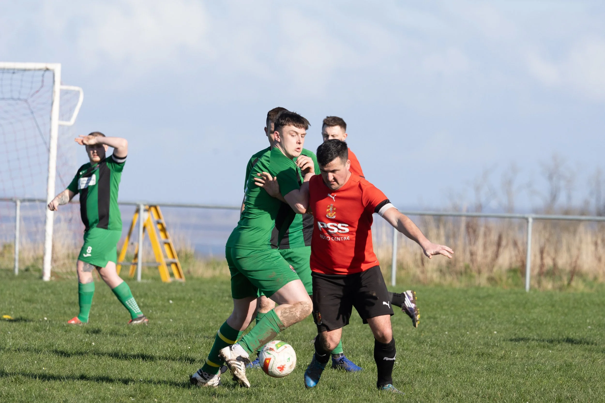 Soccer players in green and red uniforms competing for the ball on a grassy field with a goalpost in the background.