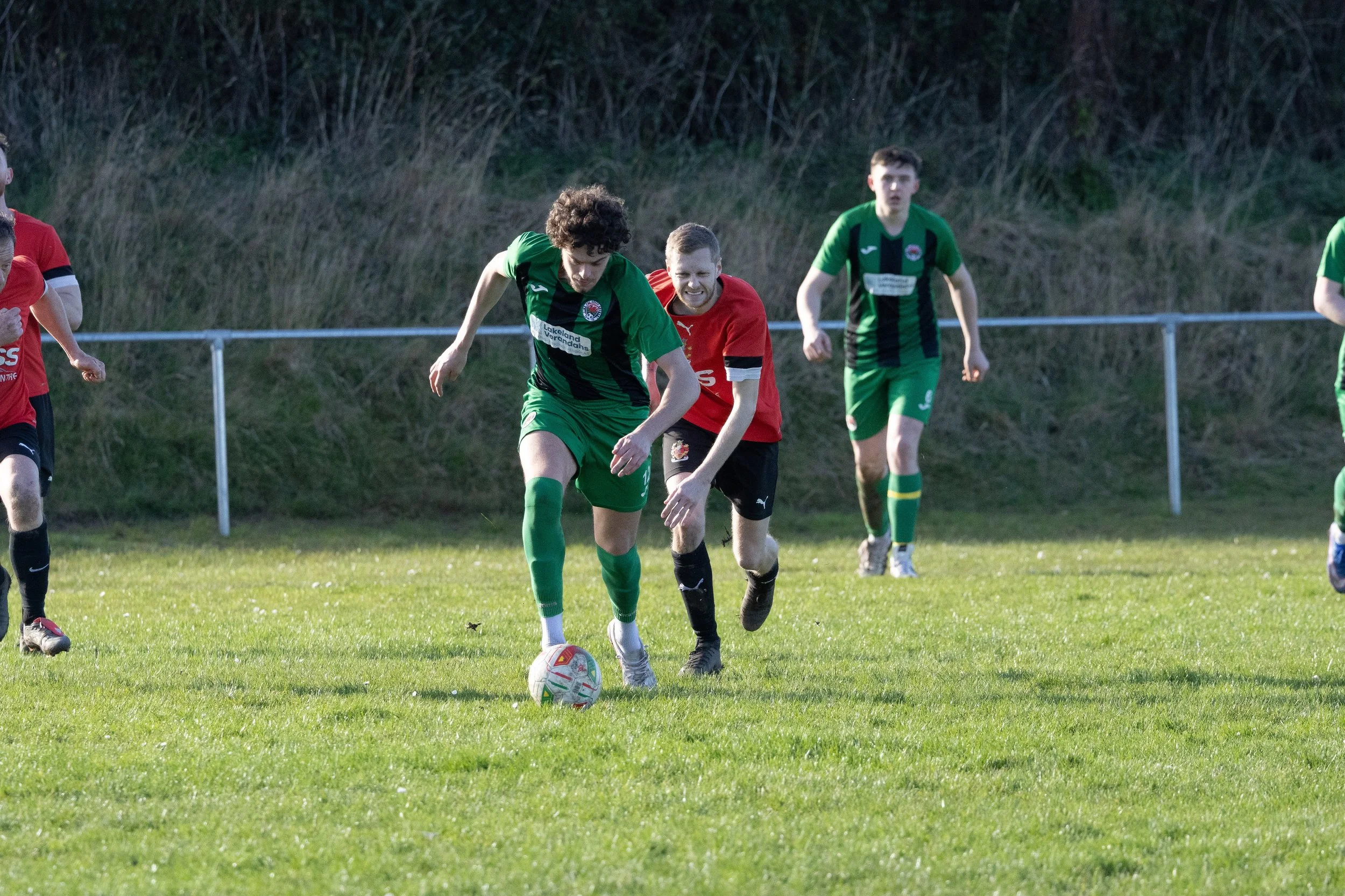Soccer players competing for the ball on a grassy field, with some players in green uniforms and others in red.