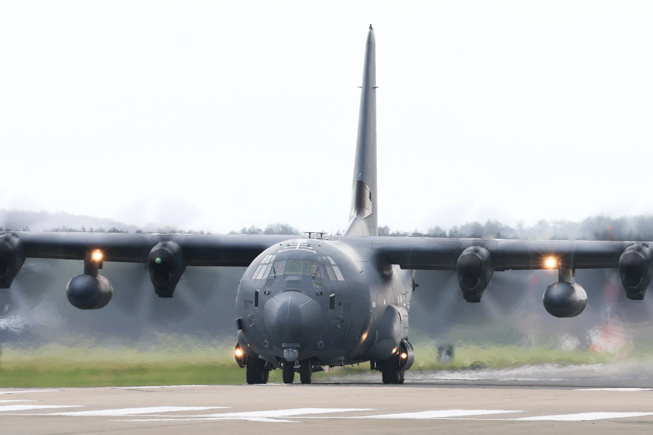A military cargo plane landing on an airstrip, with smoke and spray from the tires and engines. Overcast sky in the background.