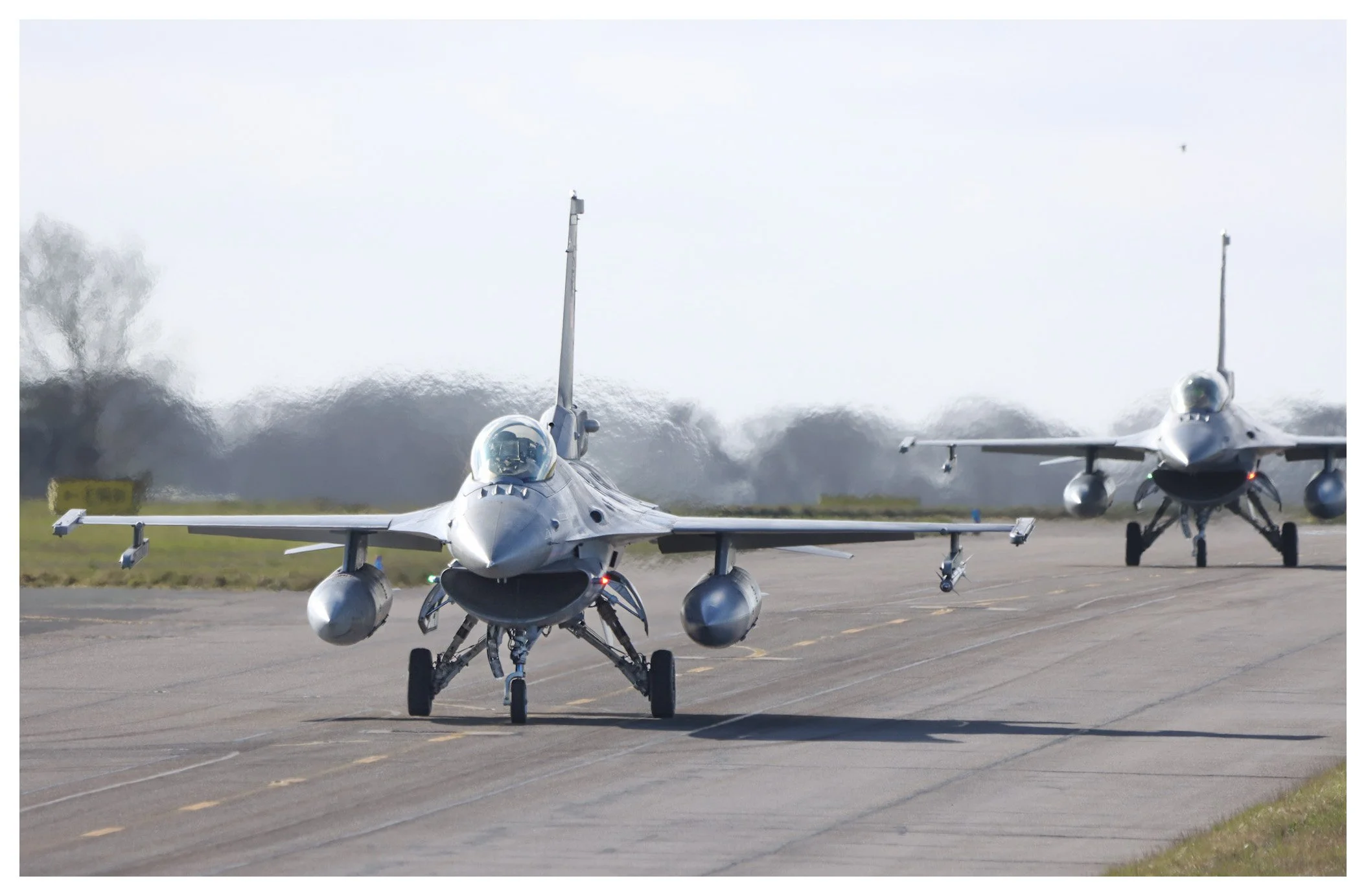 Two fighter jets on the runway during daytime, one in front and one in the background, with trees and open sky in the distance.