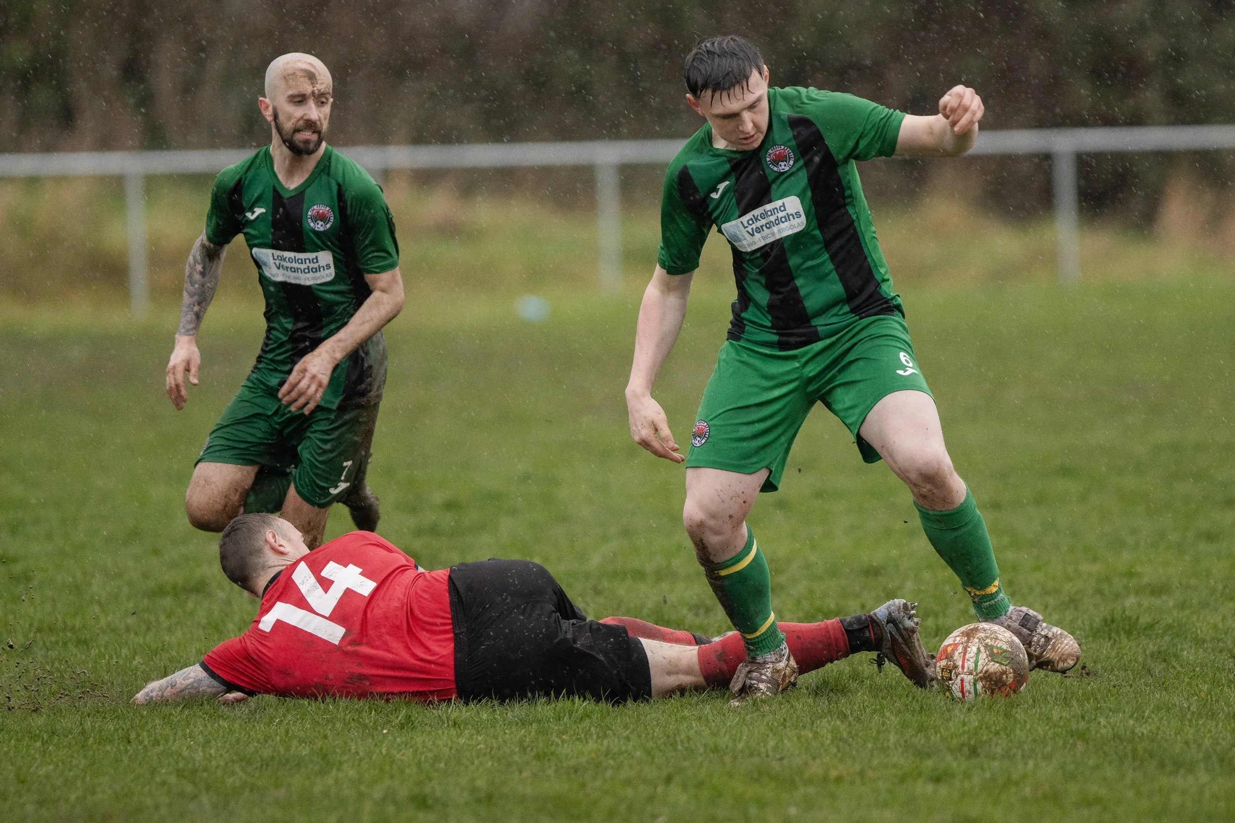 Two soccer players in green and black jerseys are attempting to get the soccer ball from a player in a red jersey and black shorts who is on the ground, during a match in rainy weather. The player in red is lying on the grass with the number 14 visib