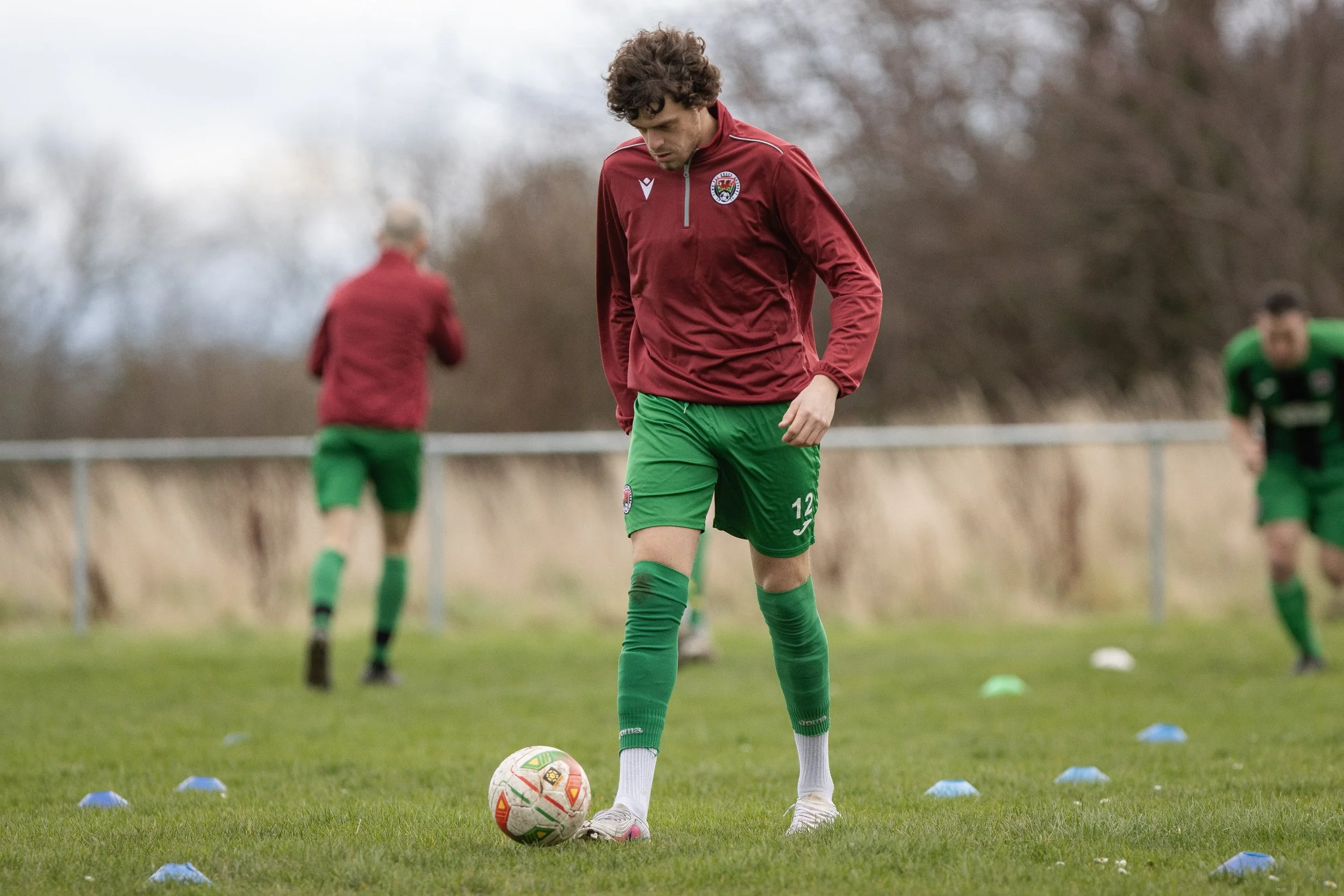 A soccer player in a red and green jersey stands on a grass field, looking down at a soccer ball, with other players training in the background.