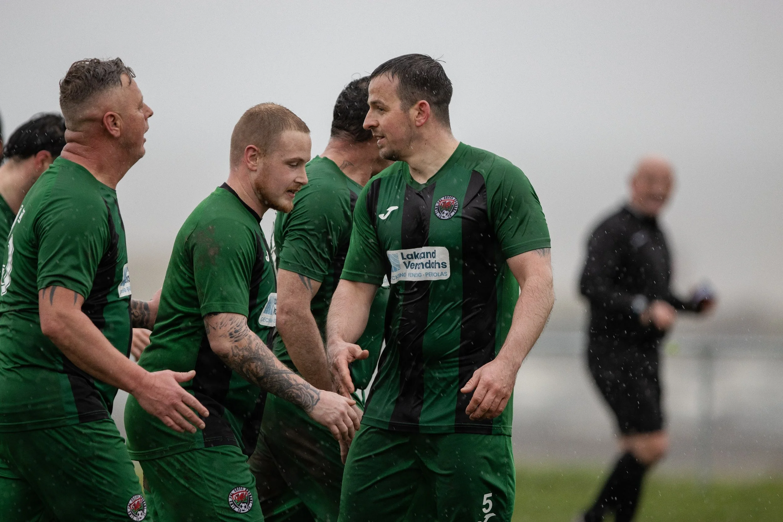 Soccer players in green jerseys celebrating on a rainy field, with one player holding hands with another, and a person in black with a camera in the background.