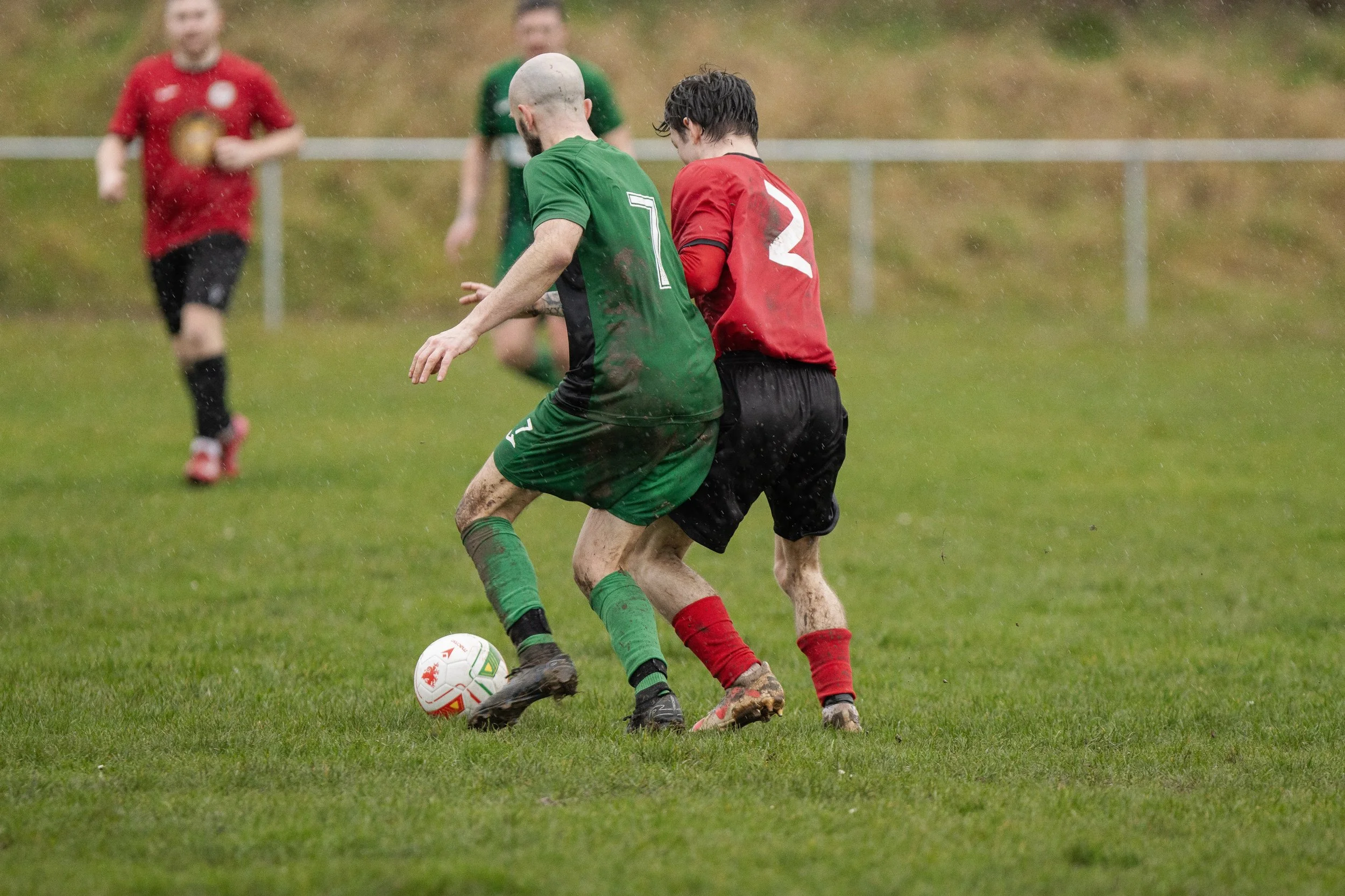 Two soccer players in green and red jerseys competing for the ball on a grassy field in the rain, with other players in the background.