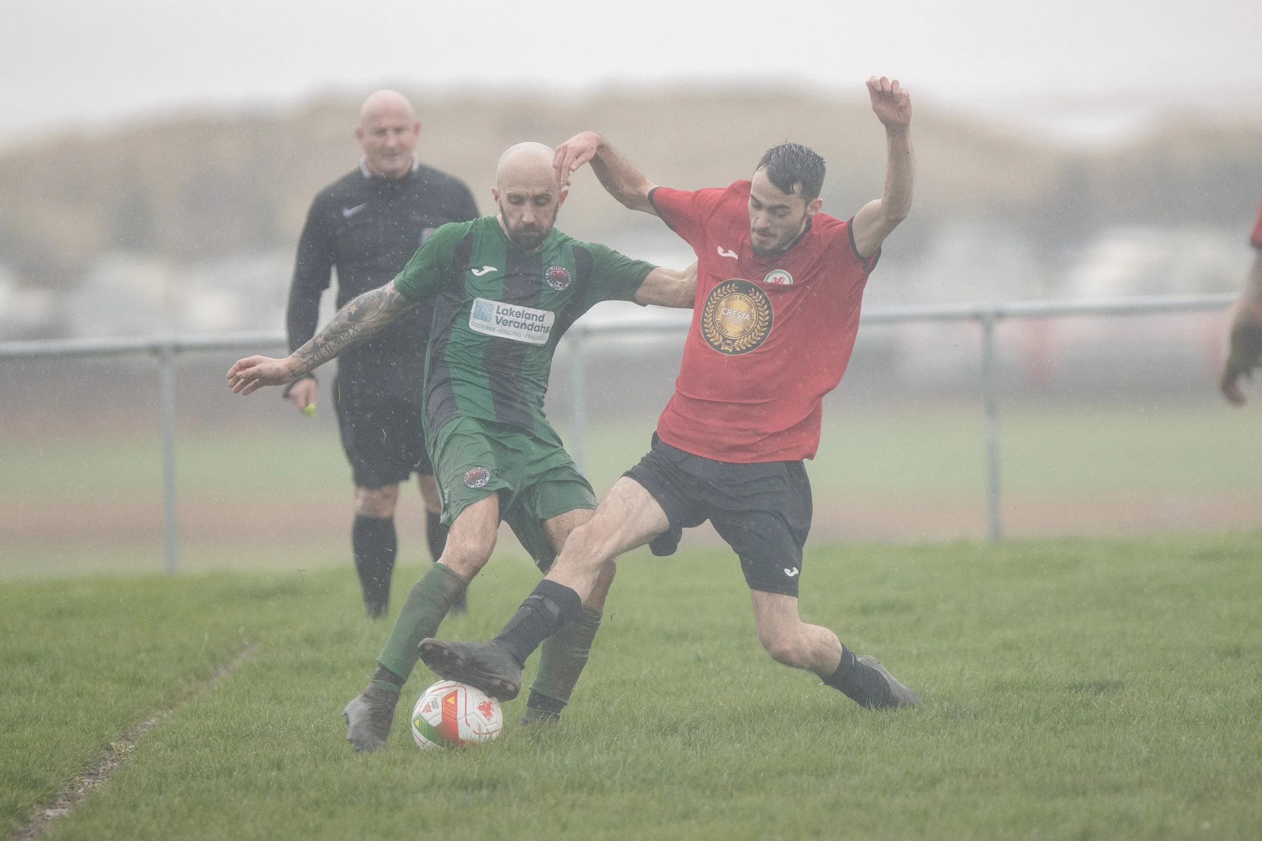 Two soccer players in green and red jerseys competing for the ball in the rain, with a referee in black in the background.