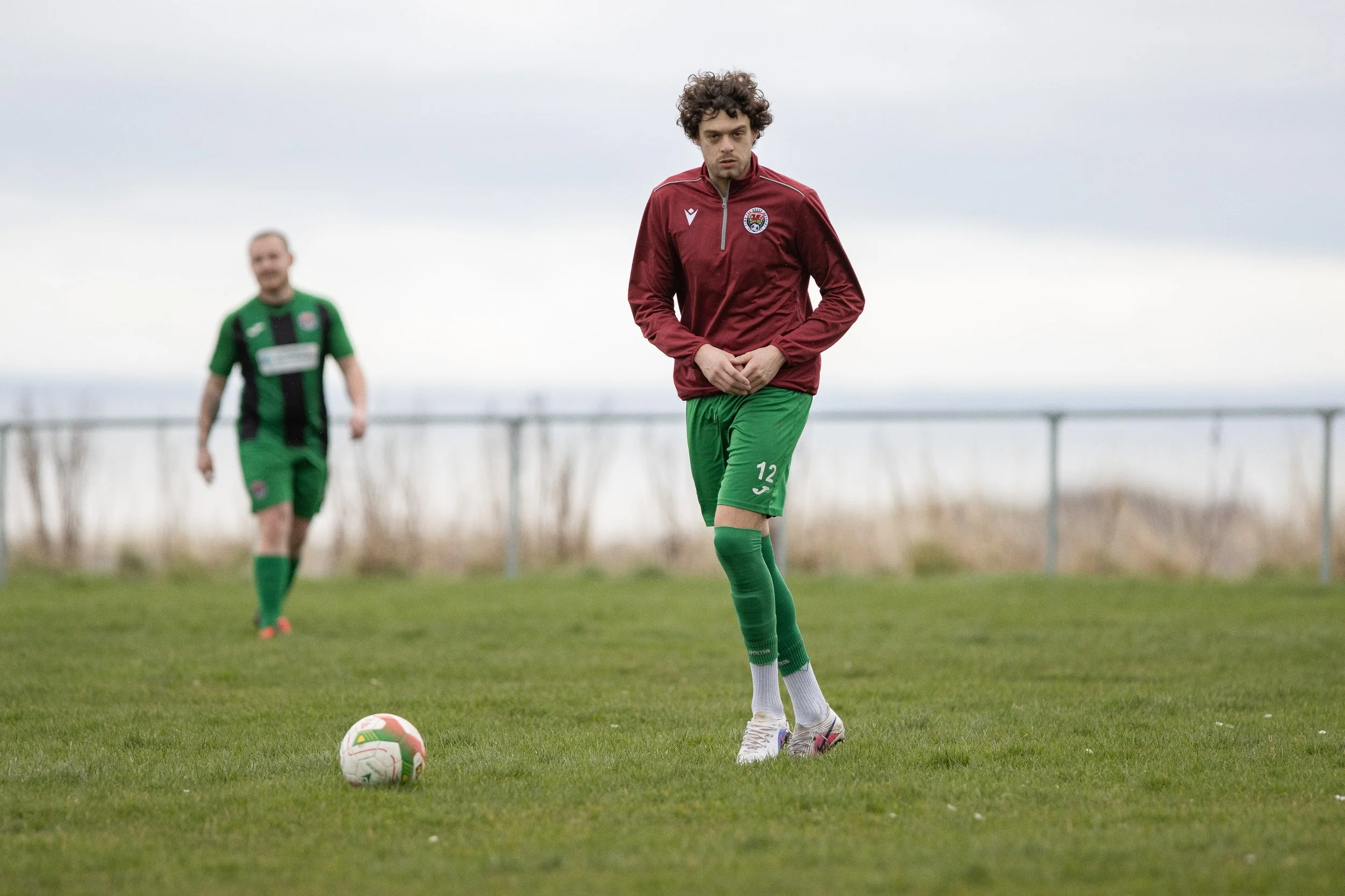 A soccer player standing on a field with a ball at his feet, wearing a maroon jacket and green shorts, with another player in the background.