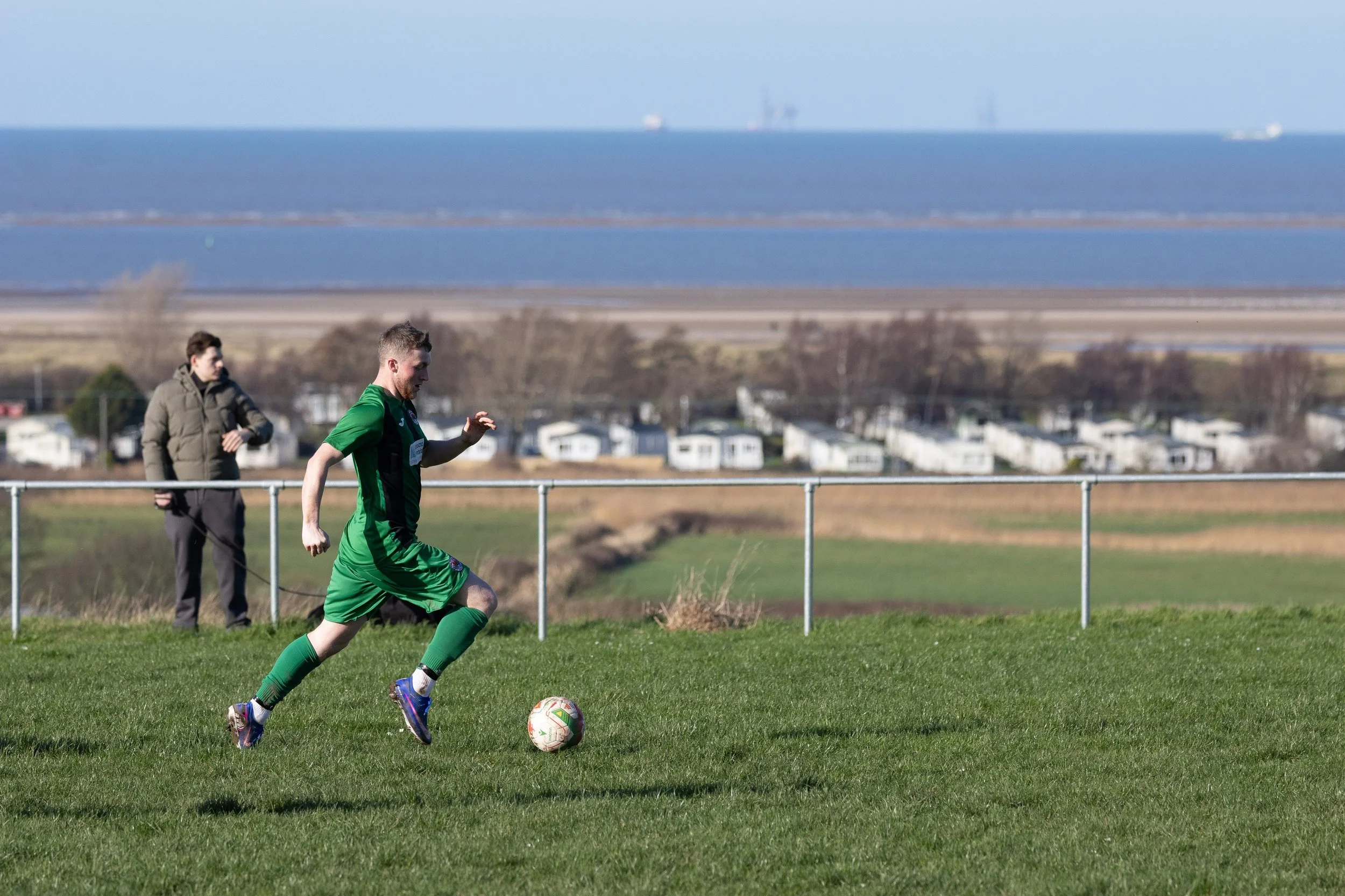 A soccer player in a green uniform running with a soccer ball on a grassy field. A person in a jacket watches in the background, and there is a chain-link fence with water and land beyond.