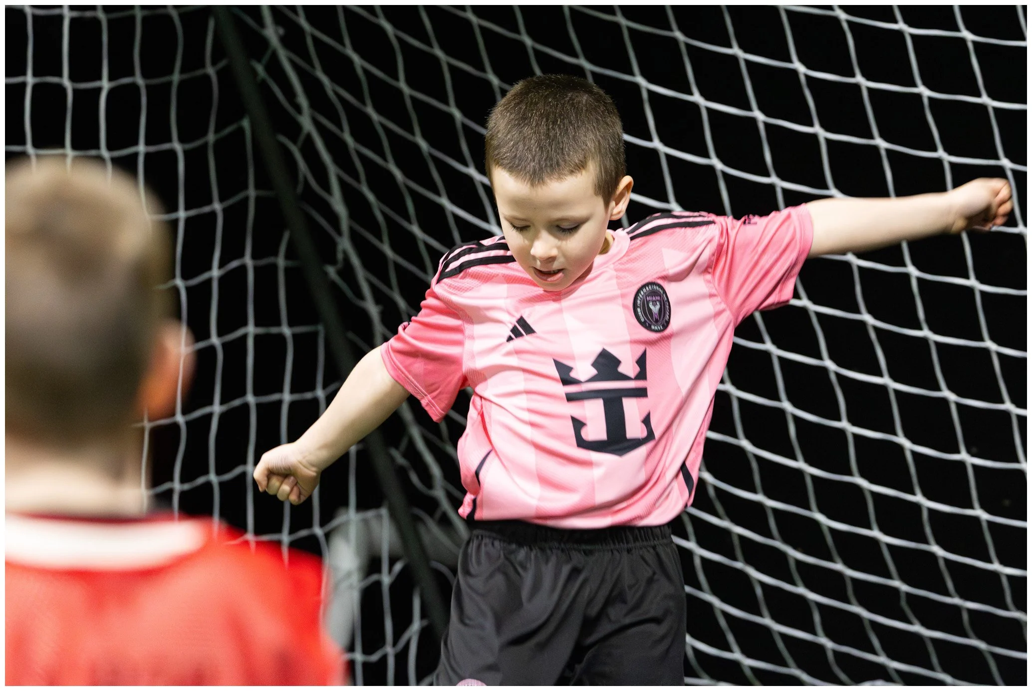 A young boy in a pink soccer jersey and black shorts is standing in front of a goal net with his arms outstretched, looking down.