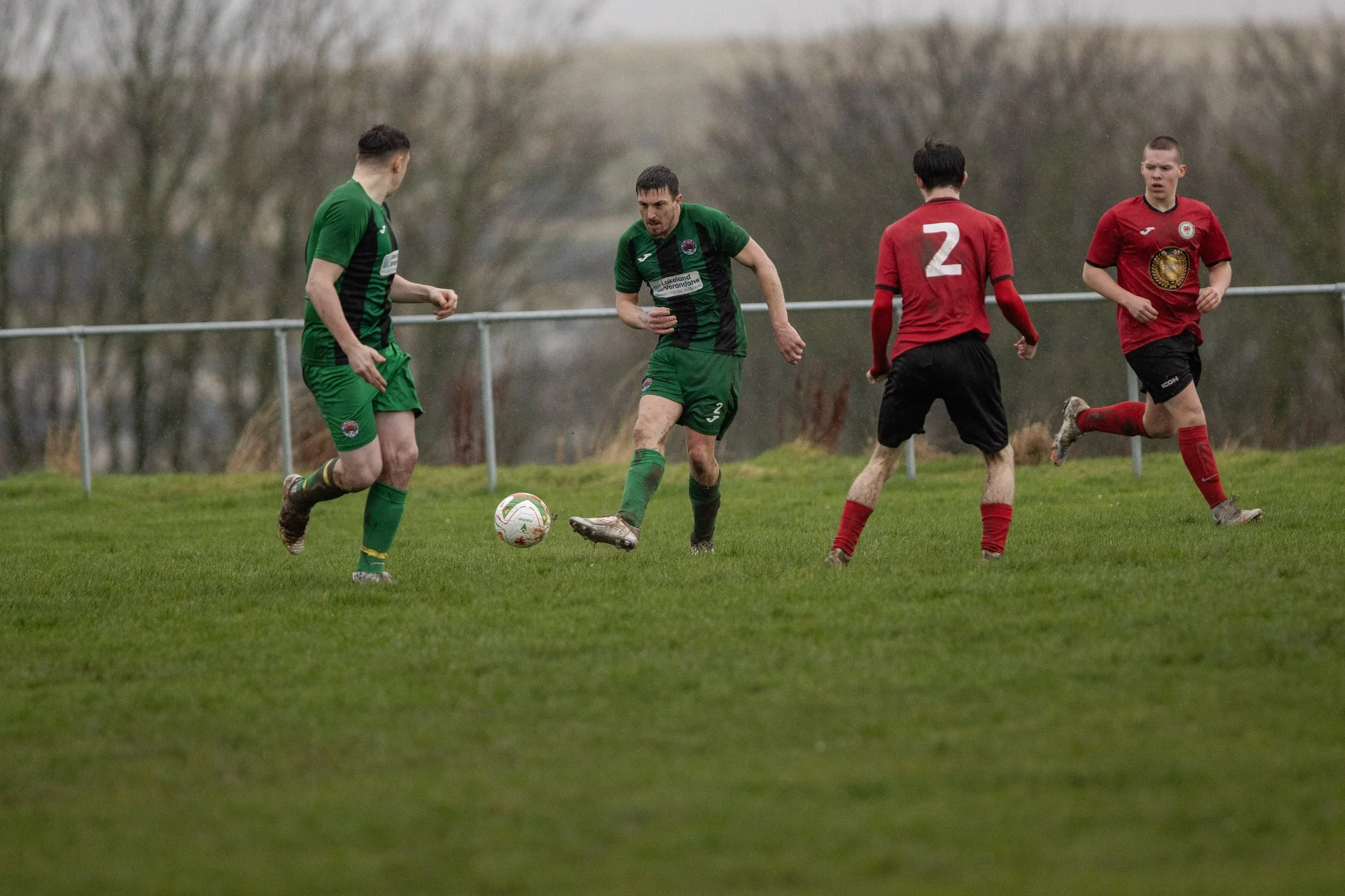 Soccer players on a grassy field during a match, with three players in red jerseys and two in green. One green player is about to kick the ball, while the others are observing or running.