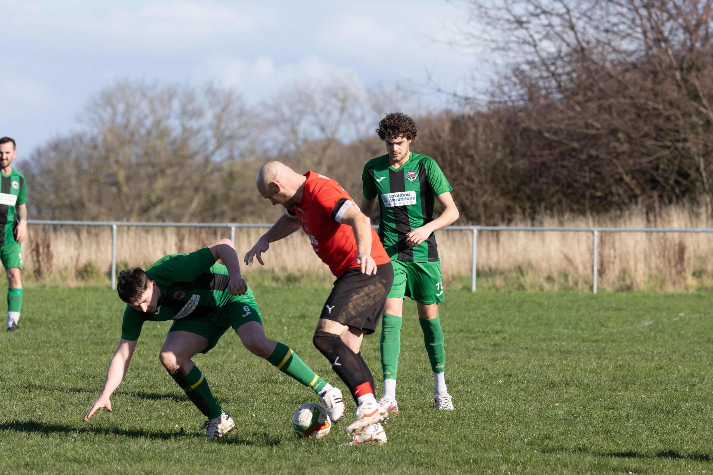 Soccer players in green and red uniforms competing for the ball on a grassy field with a fence and trees in the background.