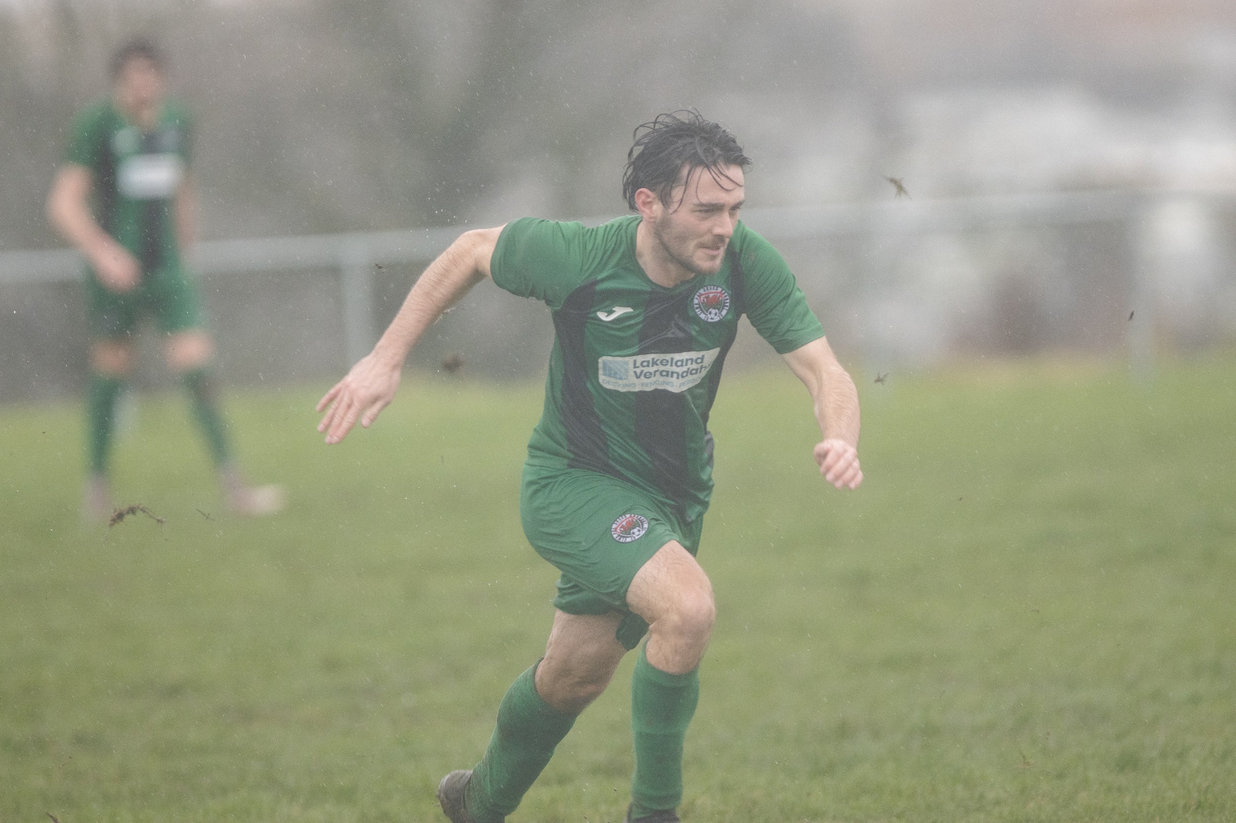 A man playing soccer in rain, wearing a green and black uniform, with another player blurred in the background.