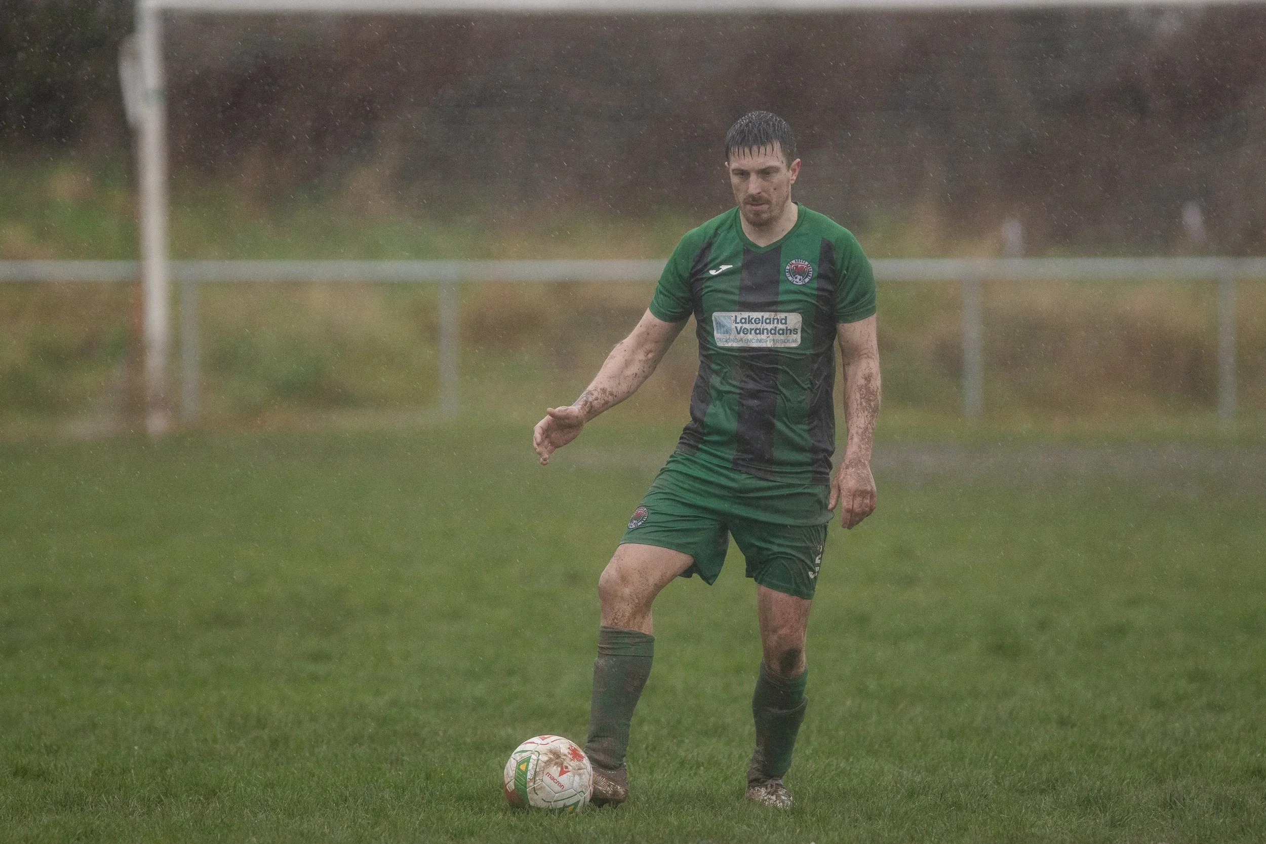 A male soccer player in a green and black uniform is playing on a field in rainy weather, with a Muddy appearance and a soccer ball near his foot.