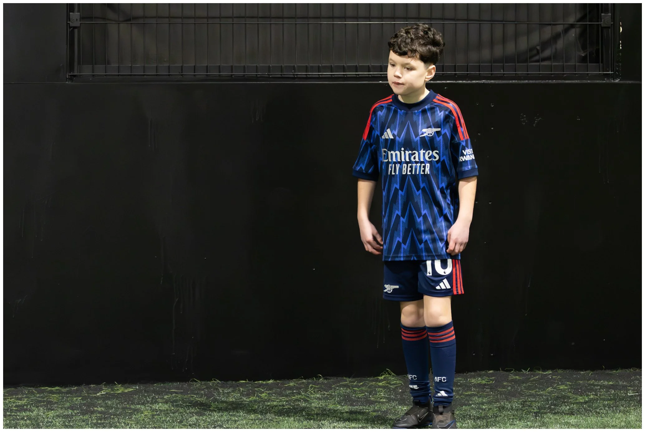 Young boy in an Arsenal football kit standing on a sports field with a black wall behind him.