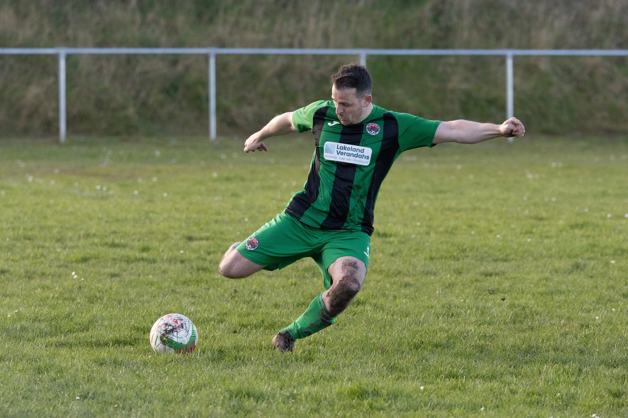 A soccer player in a green and black uniform kicks a soccer ball on a grassy field.