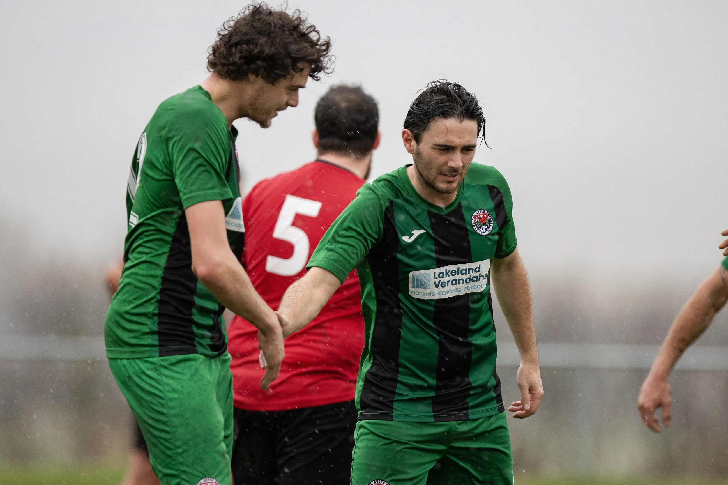 Soccer players on the field, wearing green and black uniforms, with one in a red uniform, appearing to be in a tense moment during a match.
