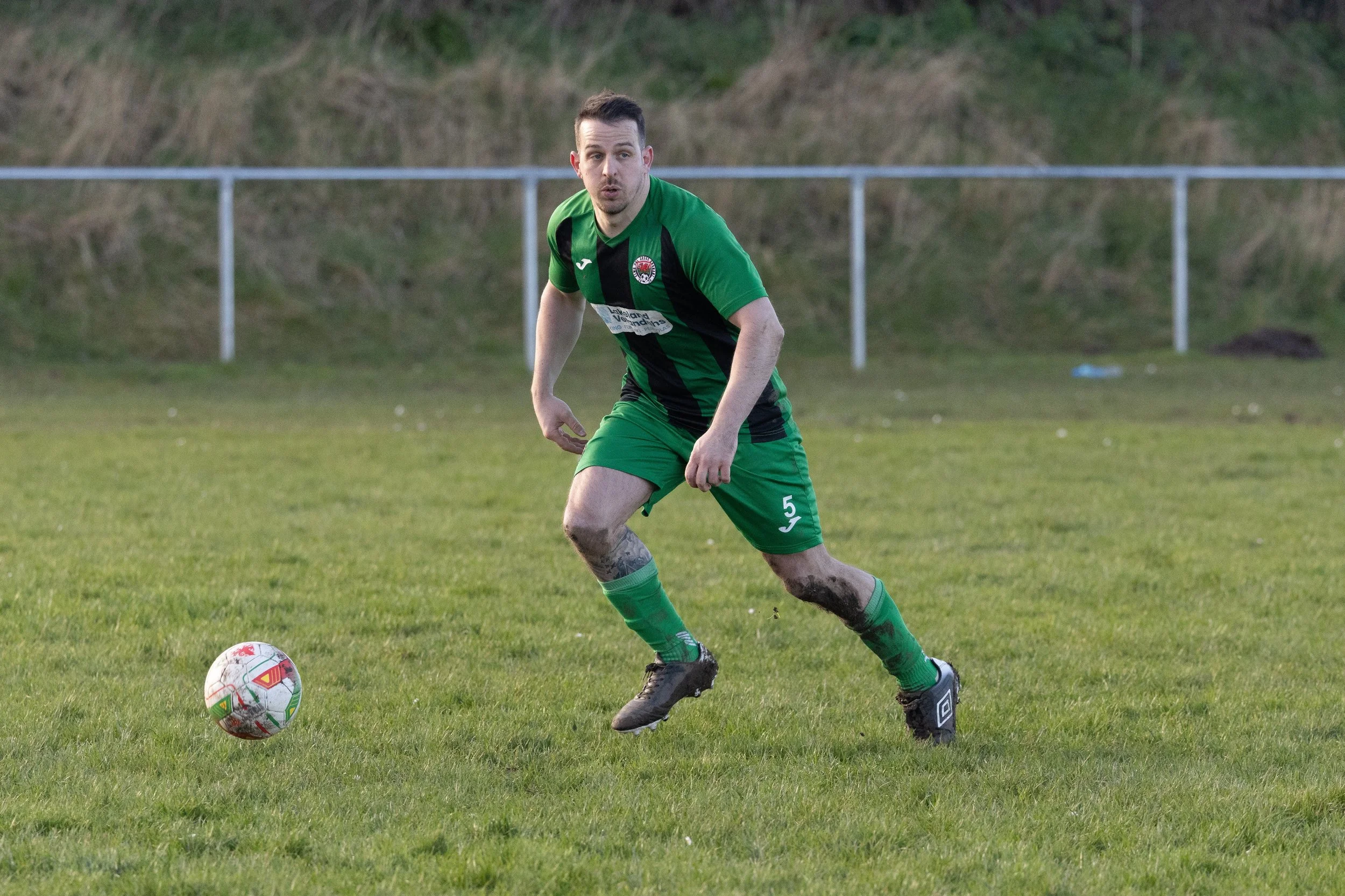 A soccer player in a green and black uniform with the number 5 is playing on a grassy field, with a soccer ball nearby, and a fence and grassy hillside in the background.