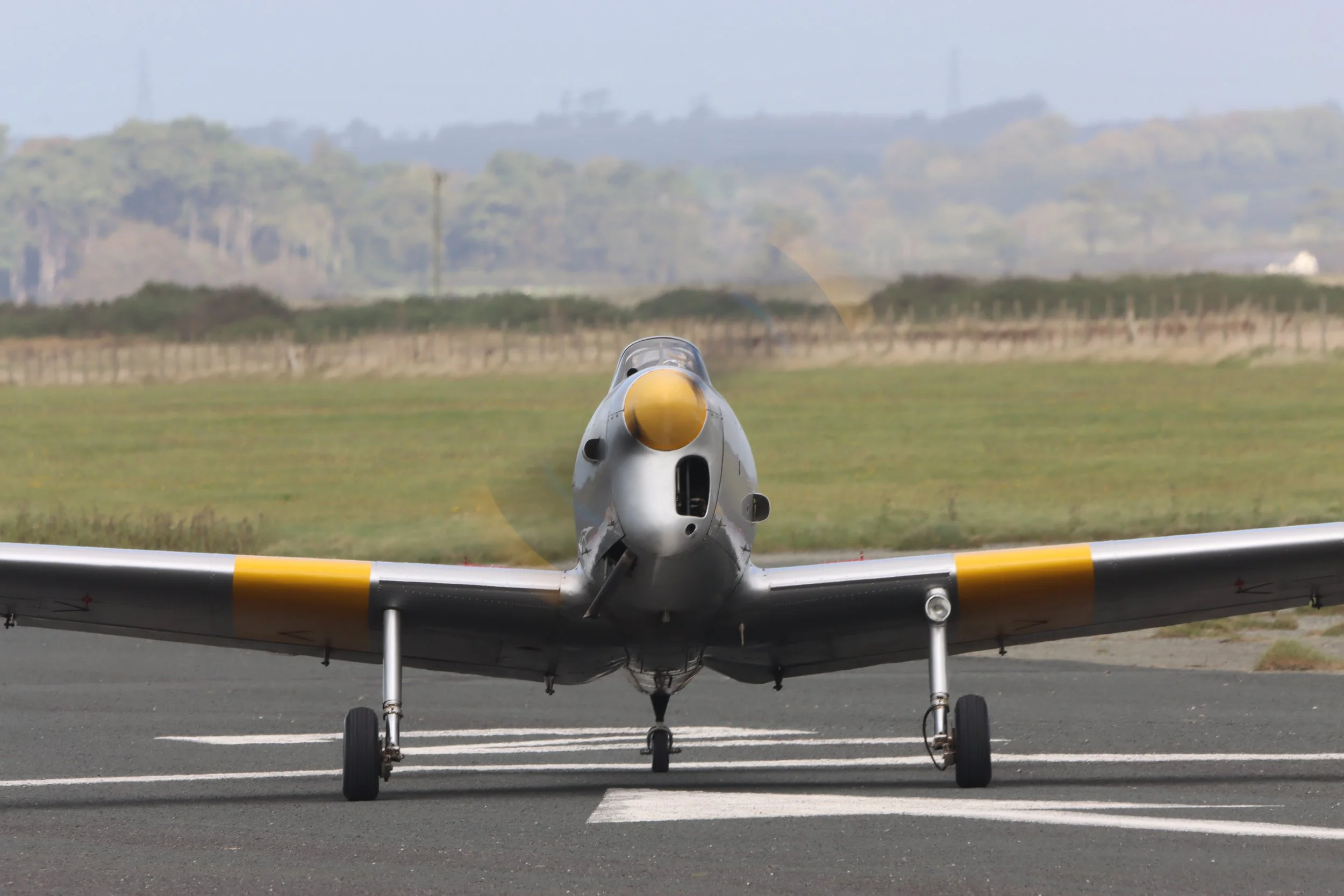 A military jet aircraft on an airport runway, facing forward, with a yellow nose cone and yellow markings on the wings, and a landscape with trees in the background.