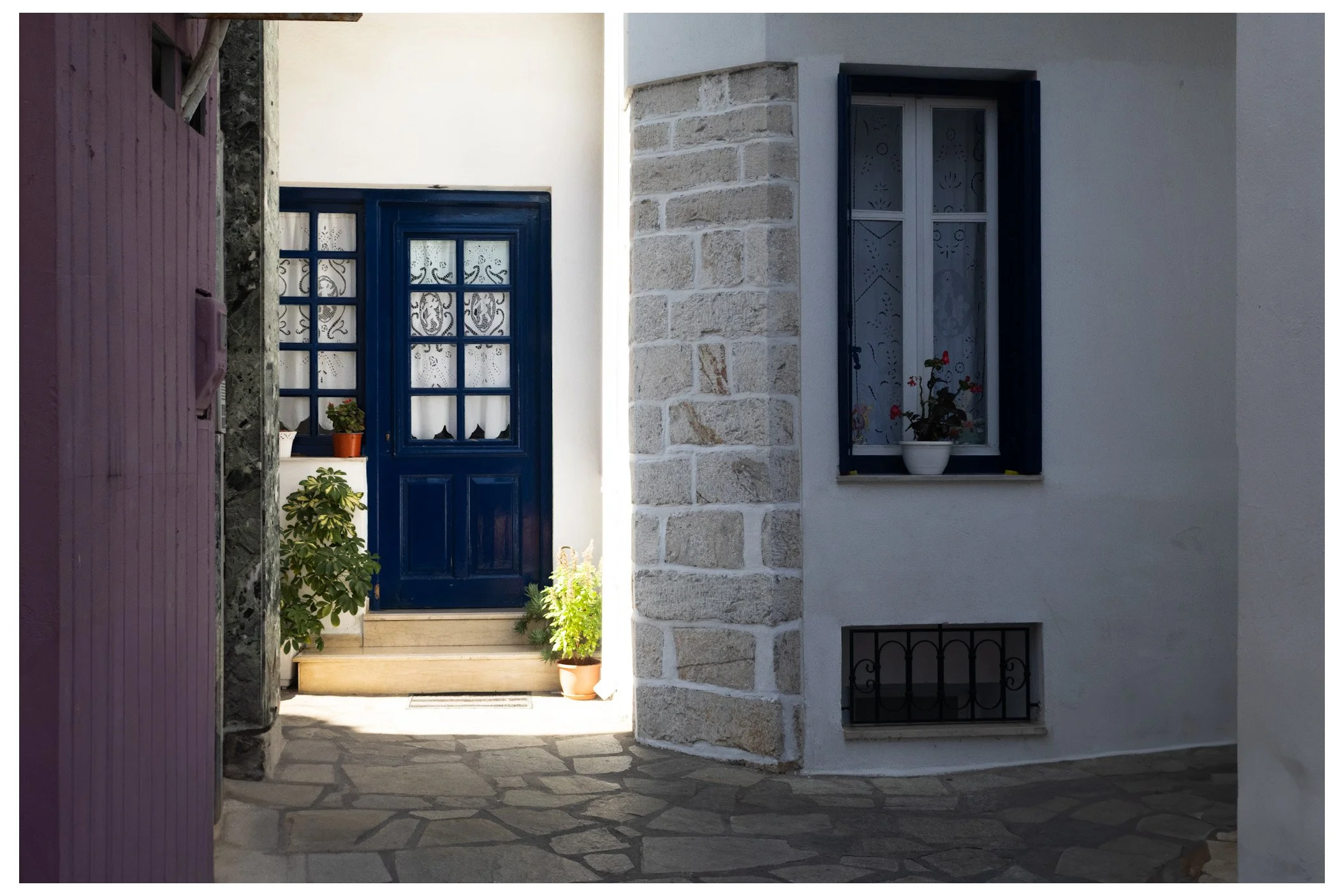 A blue door with glass panes and black decorative grille, surrounded by white walls and potted plants, next to a window with blue shutters and floral curtains, on a stone-paved pathway.