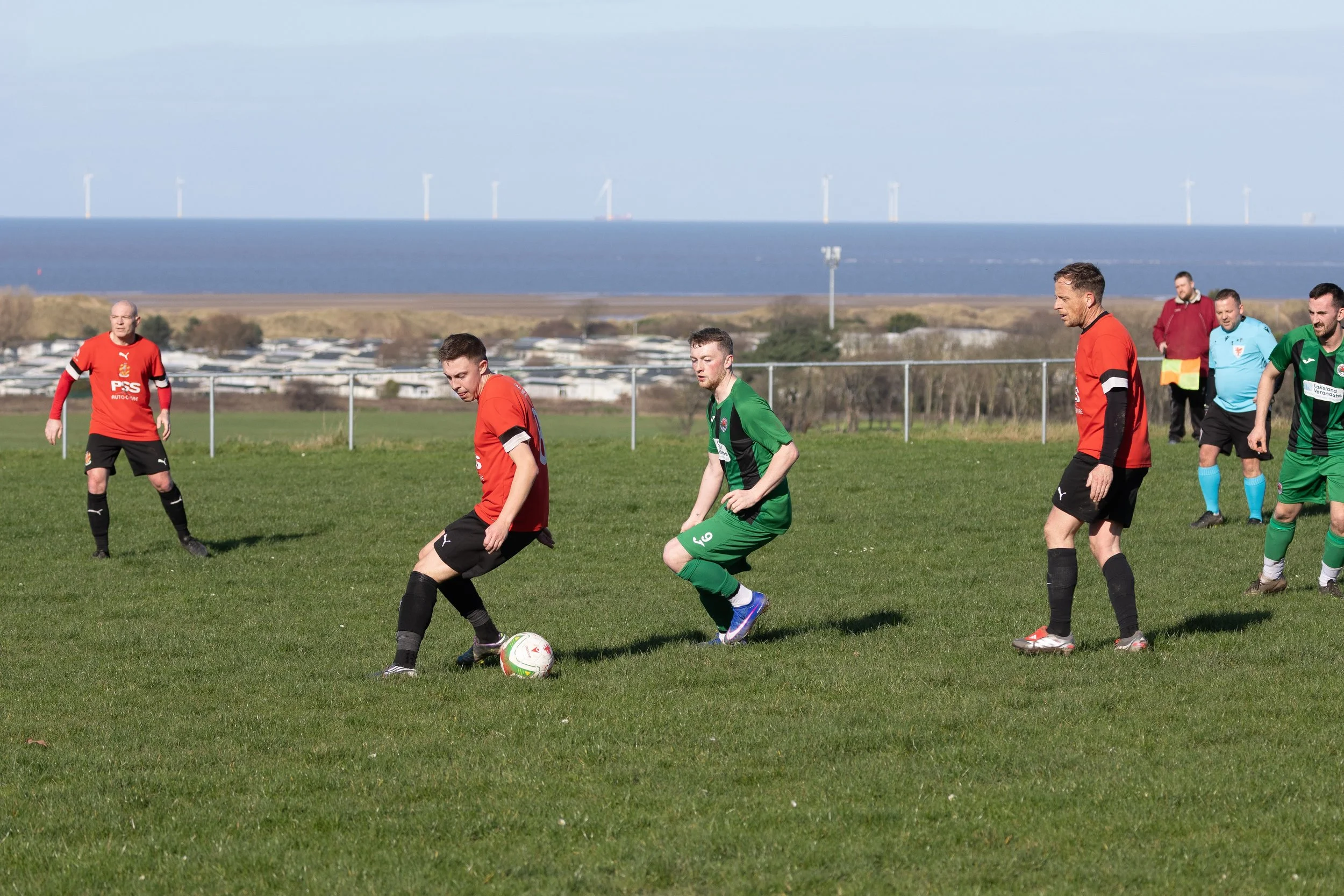 Soccer players in red and green jerseys on a grassy field near the coast, with wind turbines and a cloudy sky in the background.