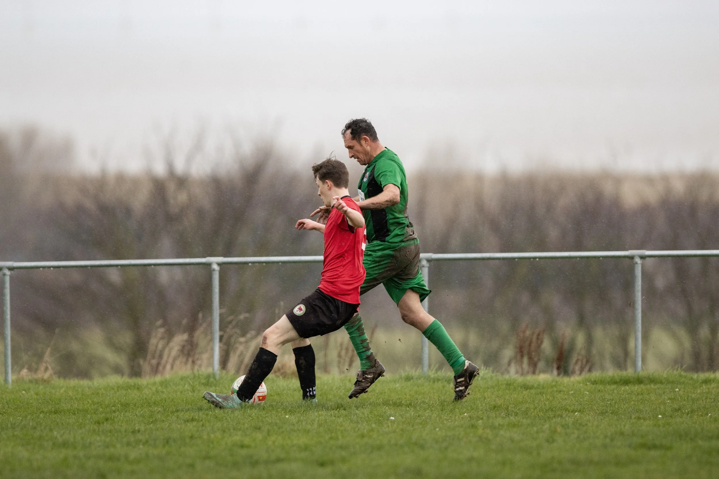 A soccer player in a red jersey and a young player in a green jersey compete for the ball on the field during a game in rainy weather.