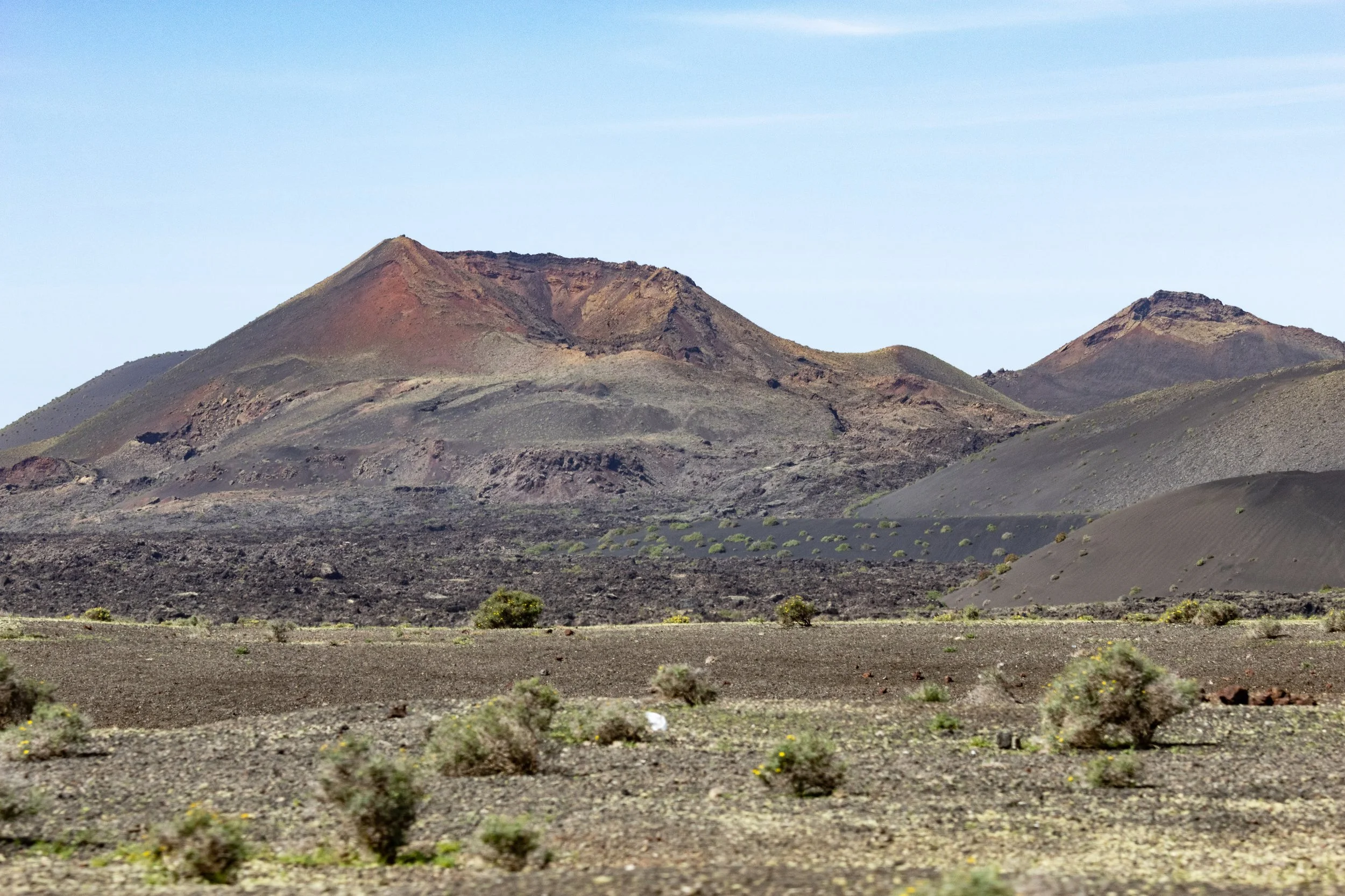 A volcanic landscape with dark, barren hills and sparse desert vegetation under a clear blue sky.