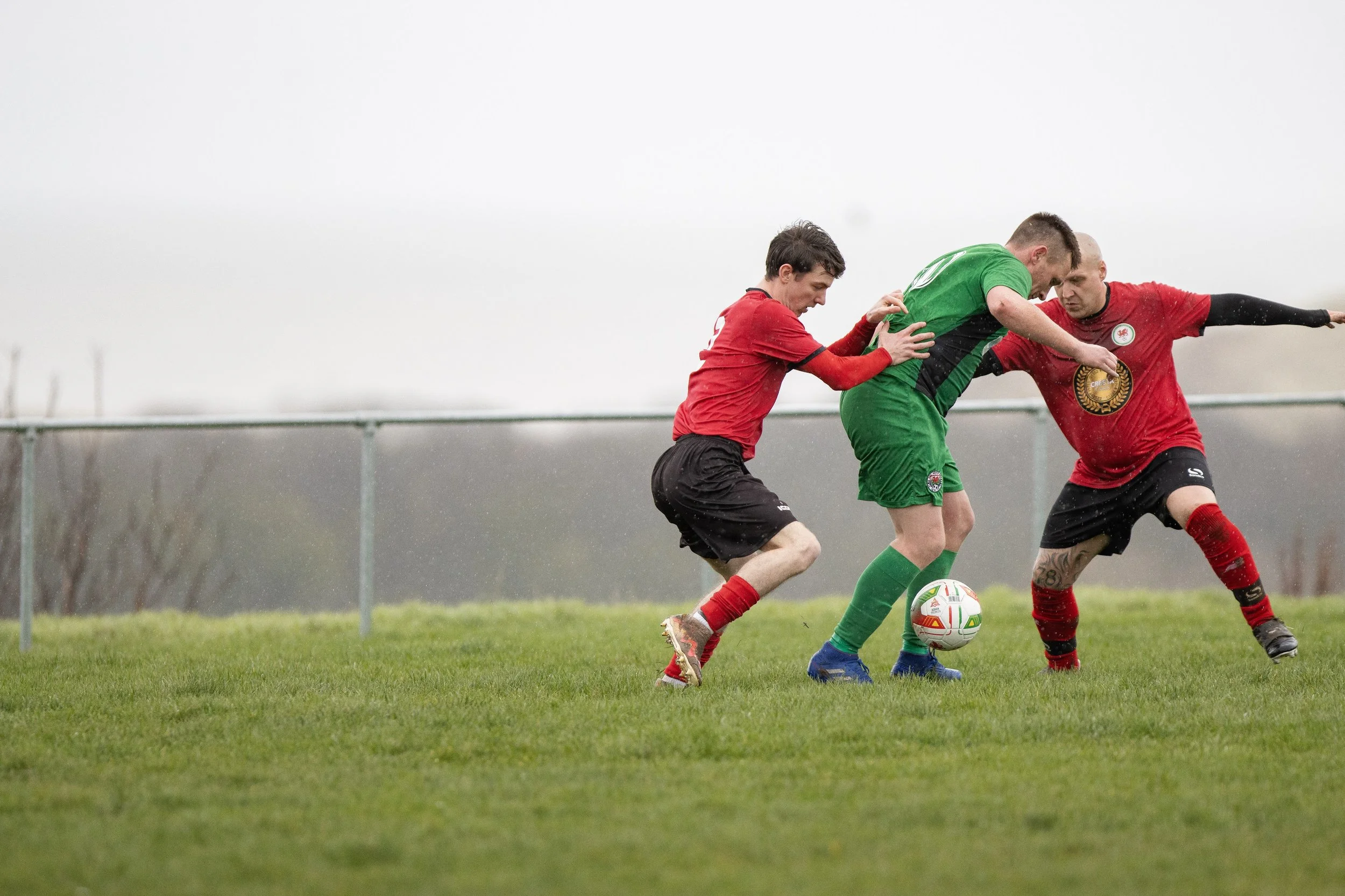 Three soccer players competing for the ball on a grassy field, with two players wearing red jerseys and one wearing a green jersey.