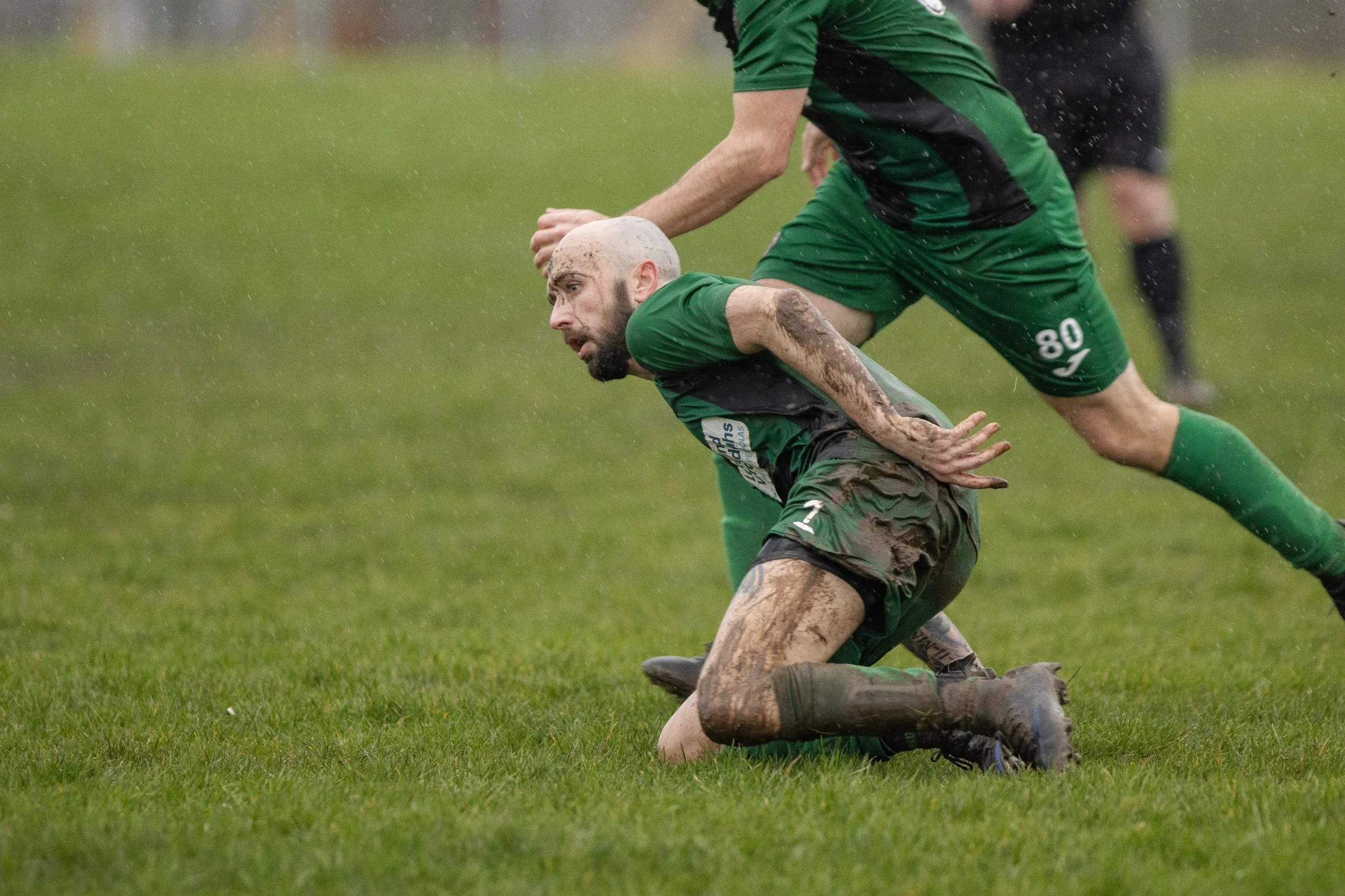 A soccer player in a green uniform kneels on the muddy grass after falling during the game, with another player partially visible above him.