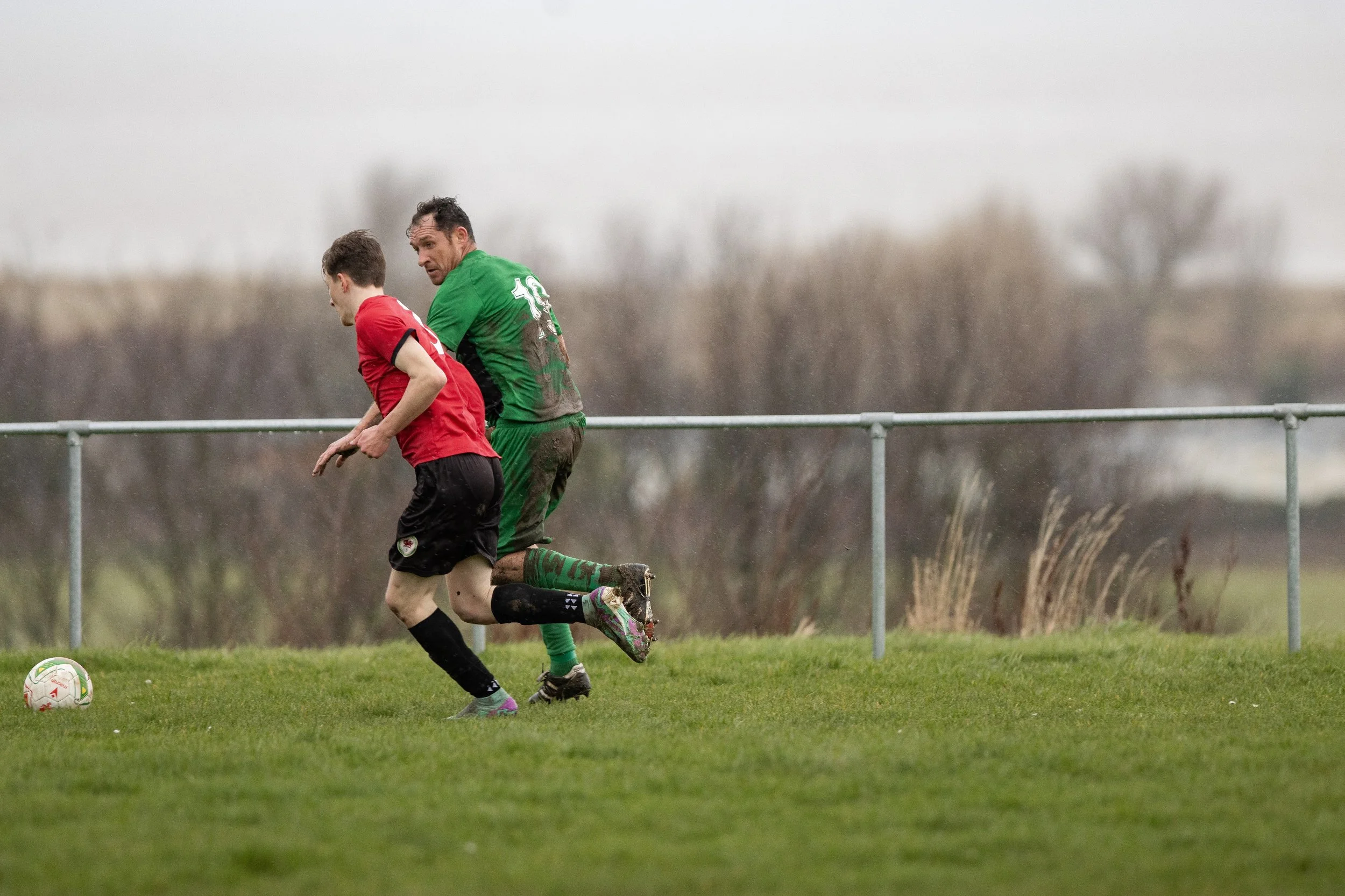 Two soccer players, one in a red jersey and the other in a green jersey, compete for control of the ball on a grassy field during a rainy day.