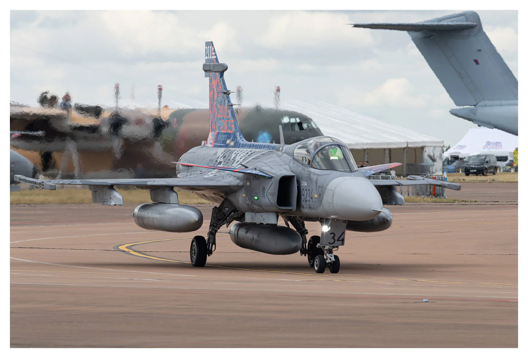 A military fighter jet taxiing on an airstrip during an airshow, with tents and other aircraft in the background.
