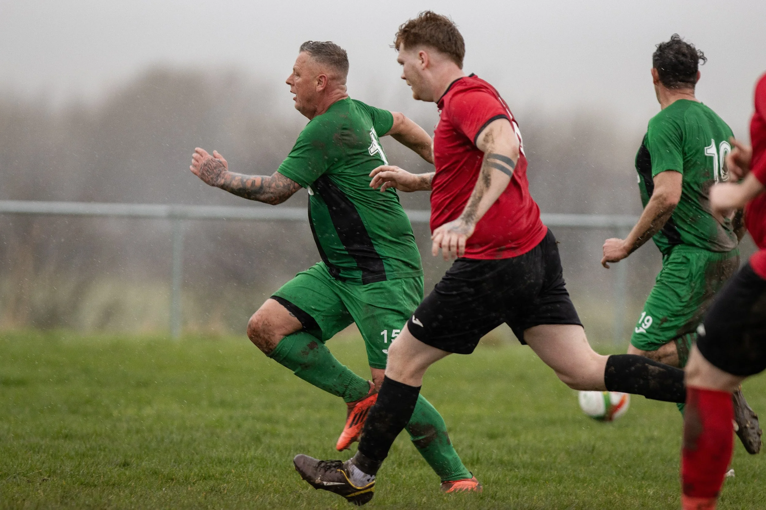 Soccer players in green and red uniforms running on a muddy field during a match in rainy weather.