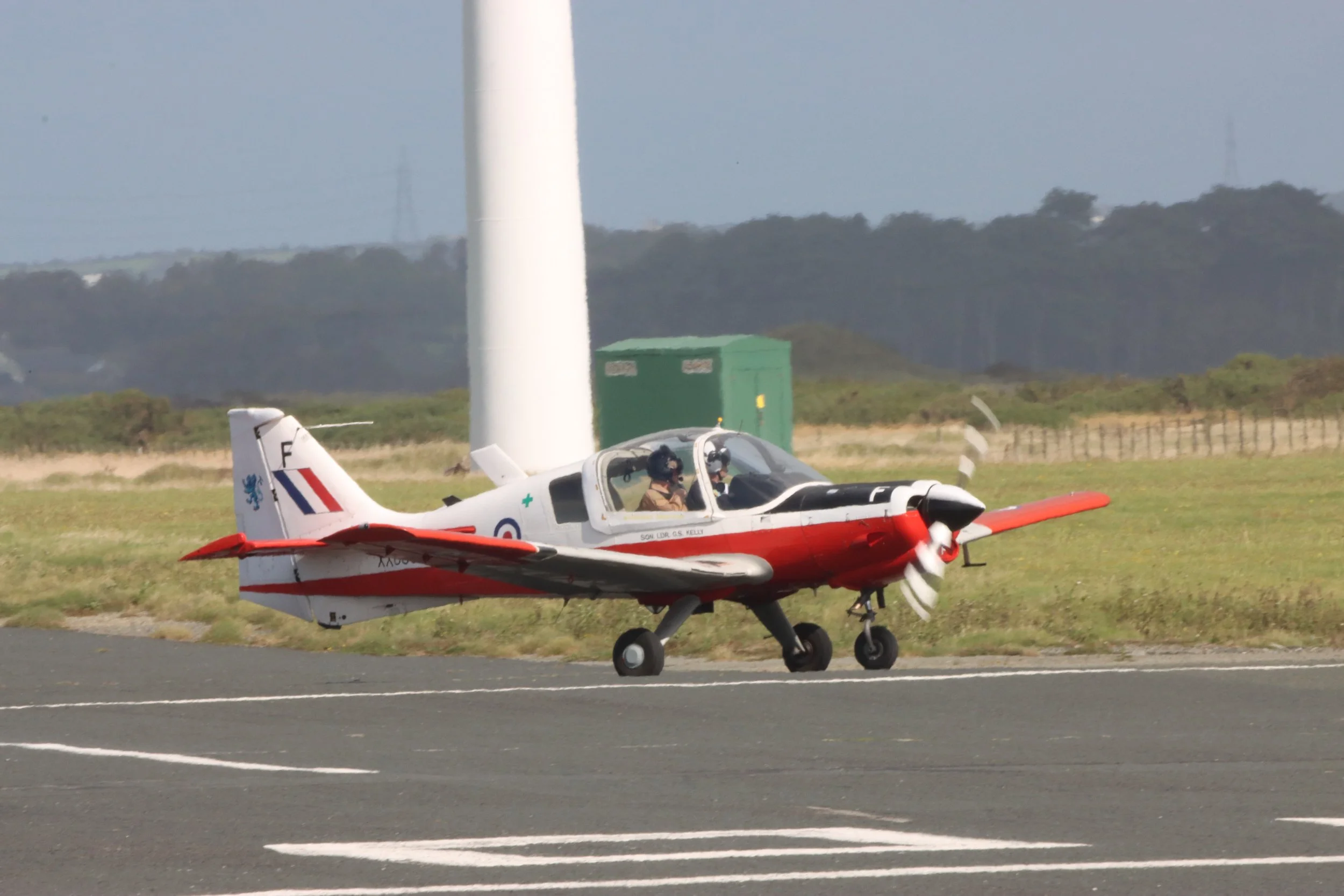 A small red and white airplane taxiing on a runway with two pilots in the cockpit, black and white striped propeller, grassy field, wind turbine, and cloudy sky in the background.