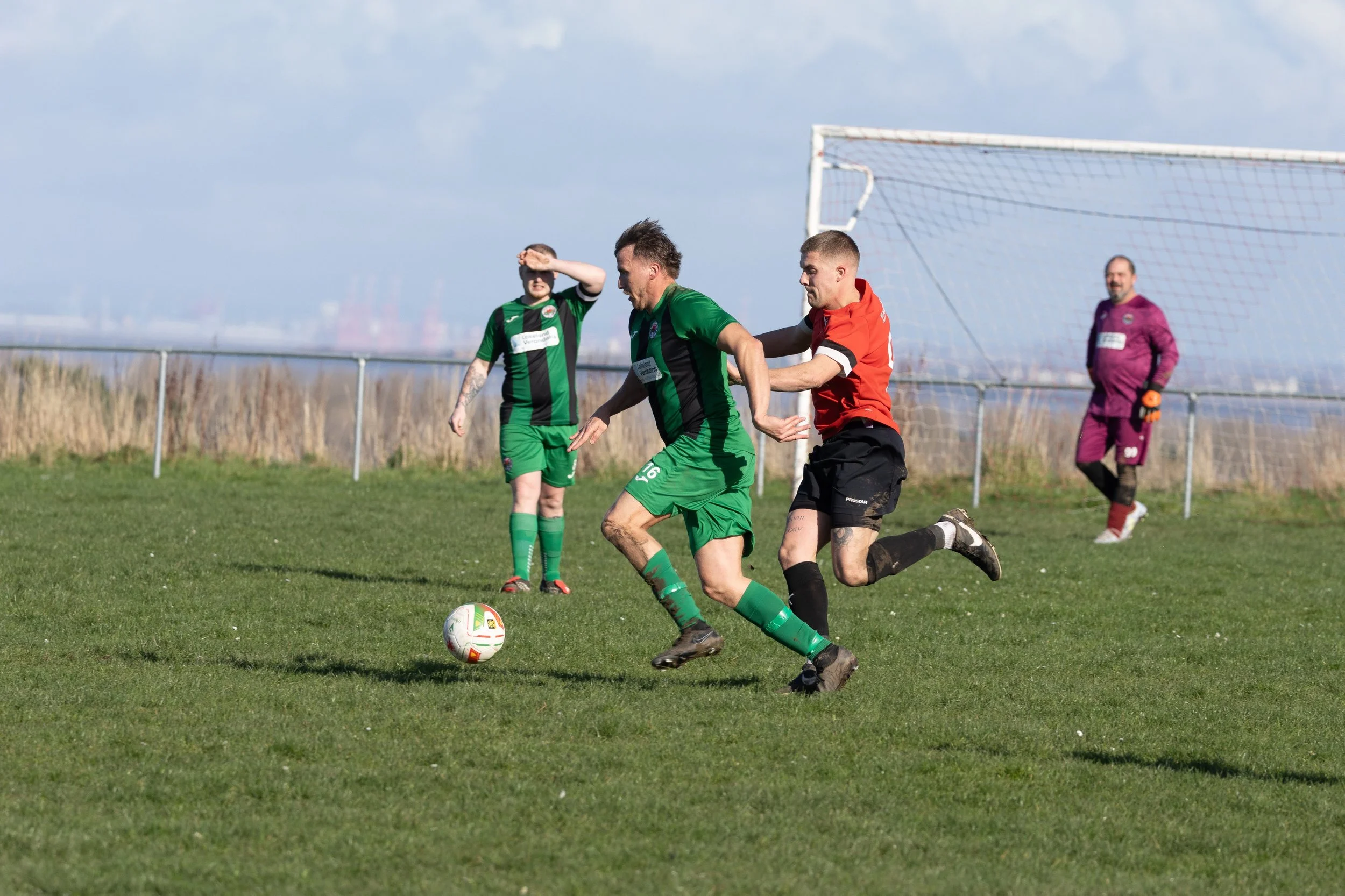 Soccer match with players in green and red jerseys; two players compete for the ball while a goalkeeper in purple stands nearby, on a grassy field under a partly cloudy sky.