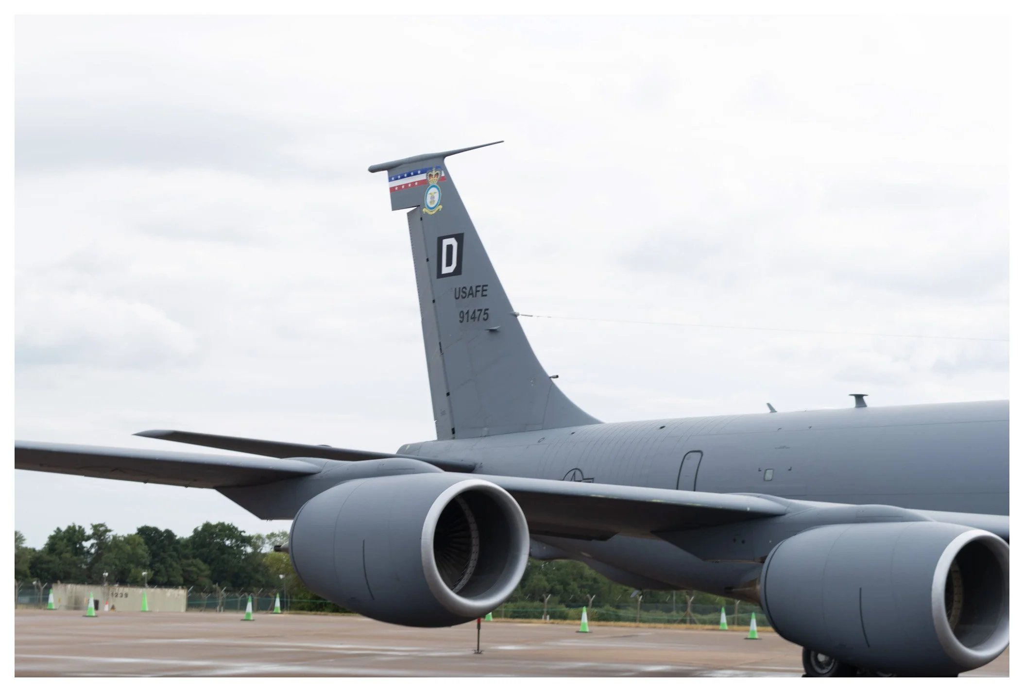 Close-up of a military aircraft, focusing on the tail and jet engines, parked on an airstrip with orange cones and a cloudy sky.