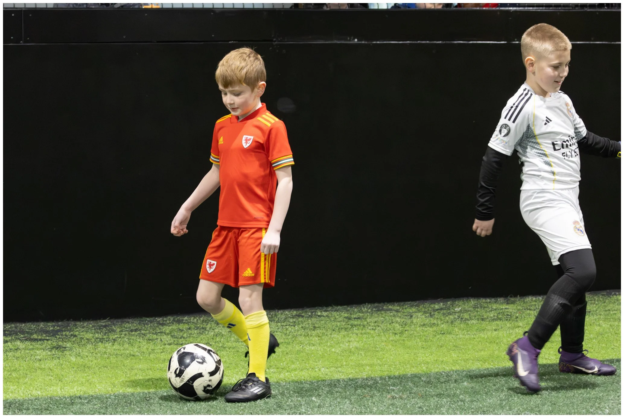 Two young boys playing soccer indoors, one in a red Welsh national team jersey and yellow socks, controlling a black and white soccer ball, the other in a white Real Madrid jersey with black sleeves and purple shoes, on an artificial grass field.