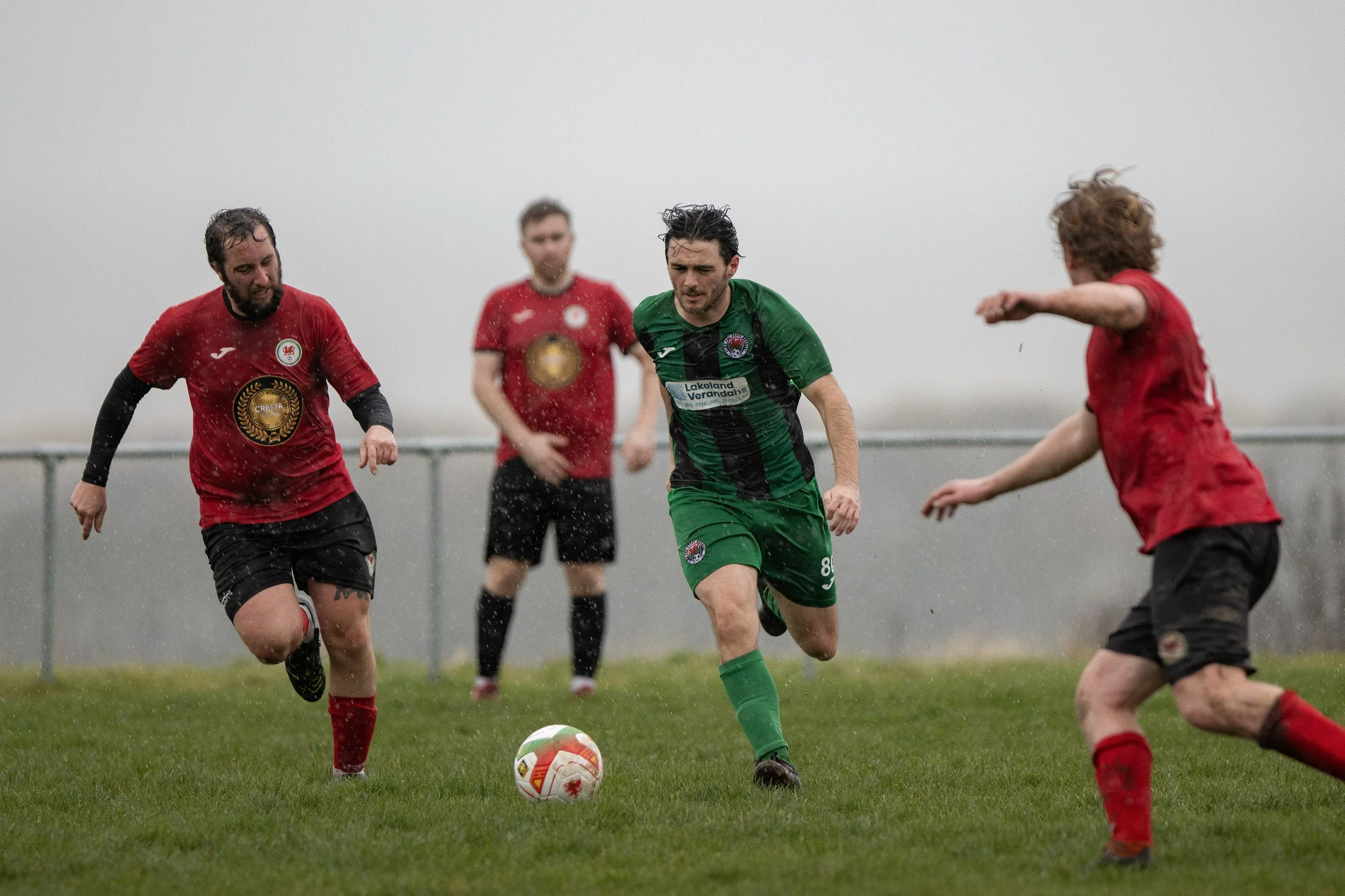 Soccer players playing in the rain on a grassy field, with one player in a green uniform dribbling the ball past two players in red uniforms.