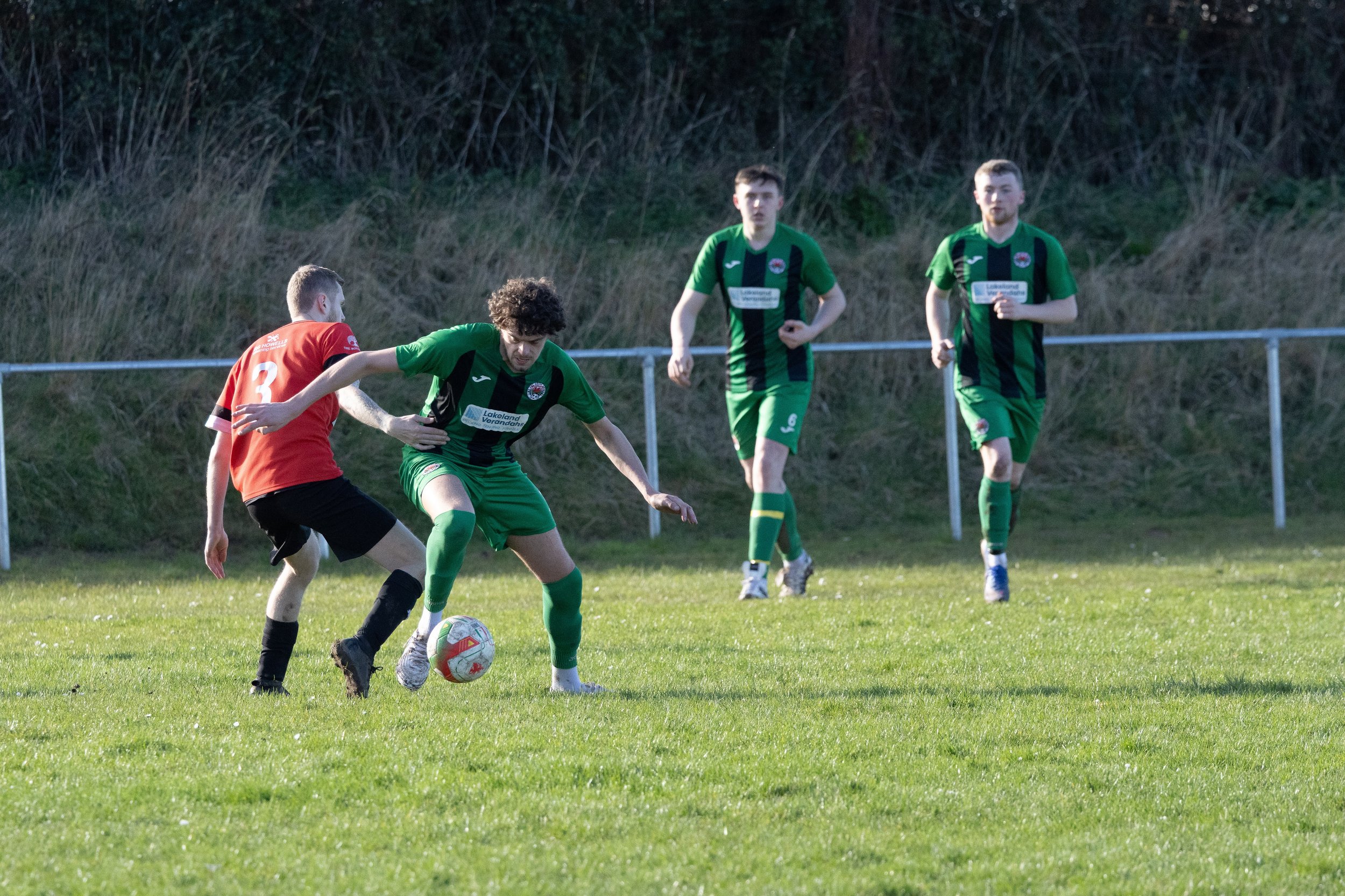 Soccer players competing for the ball during a match on a grassy field, with three players in green and one in red, with a grassy hillside in the background.