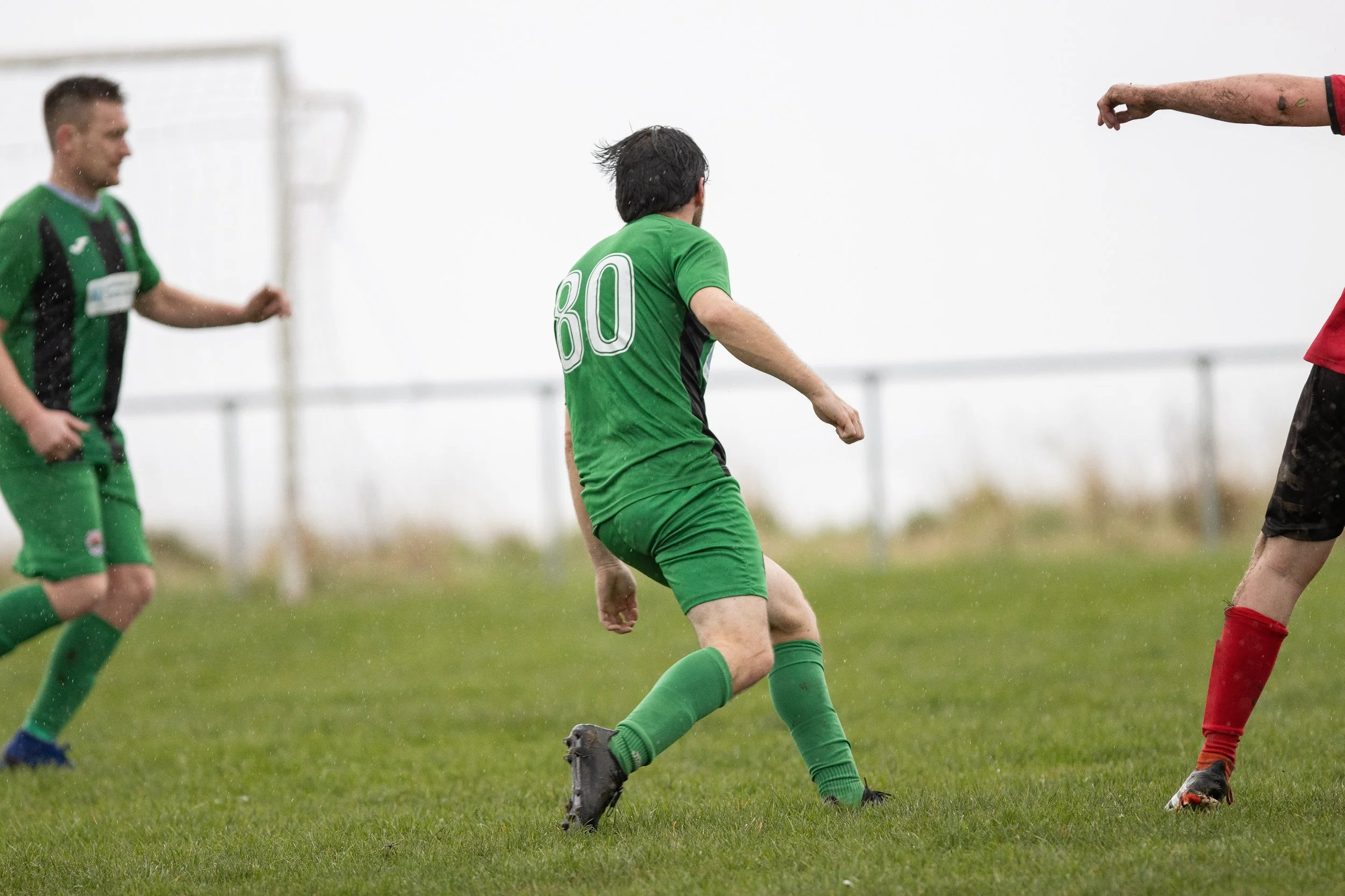 Soccer players in green and red uniforms playing on a rainy day on grassy field.