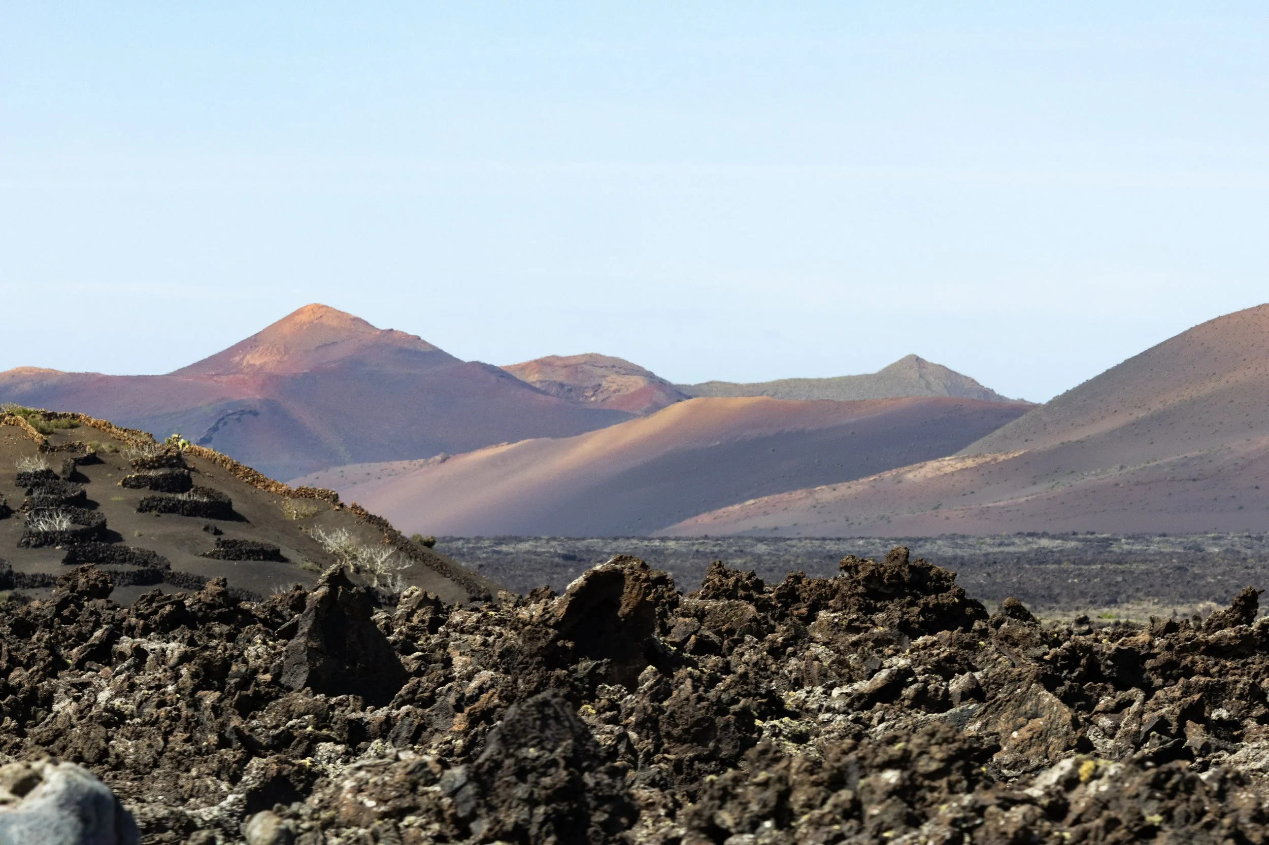 A barren volcanic landscape with dark, jagged rocks in the foreground and rolling hills and mountains in the background under a clear blue sky.