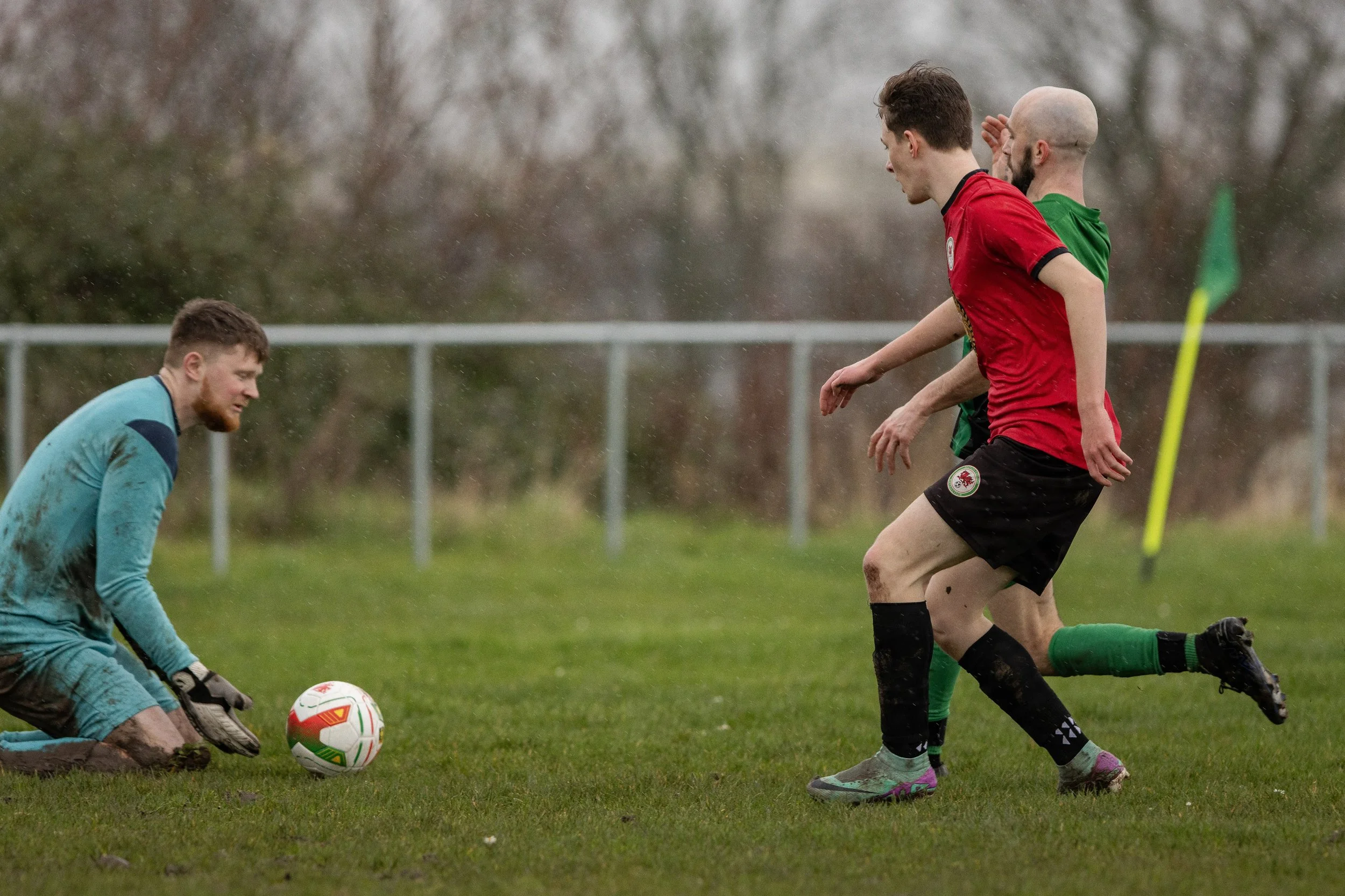 A soccer game on a rainy day with players in red and green jerseys, a goalkeeper in a light blue uniform on the ground near the goal, and two players in red and green chasing the ball near a corner flag.