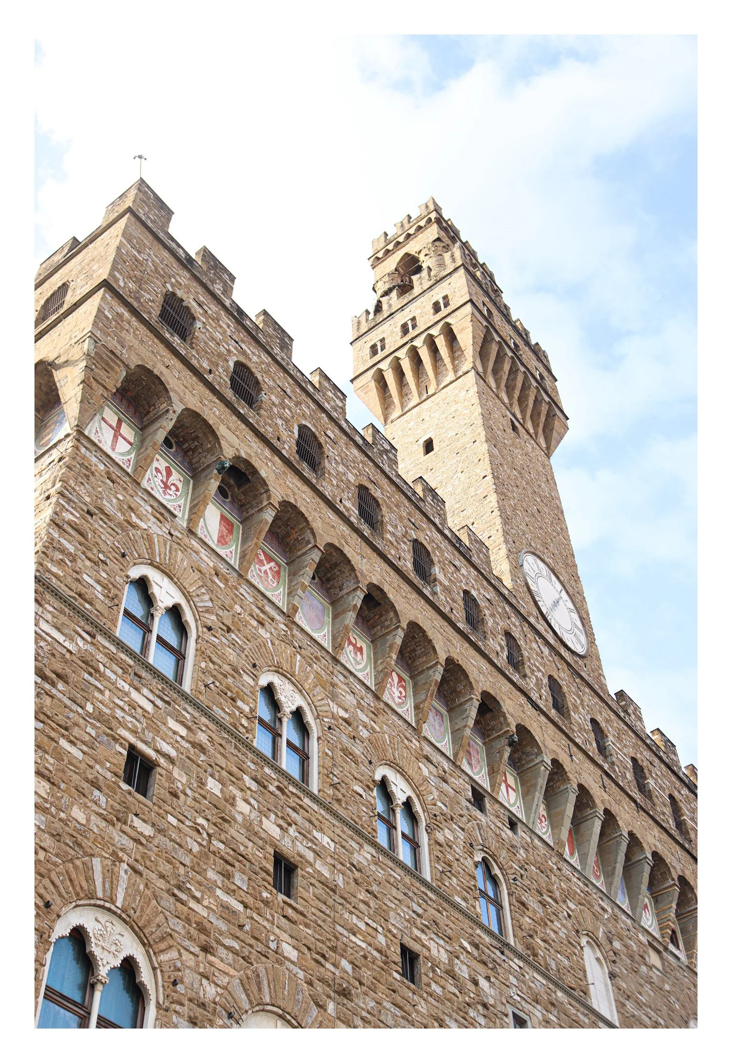 View of the historic Palazzo Vecchio in Florence, Italy, showing its stone facade, windows with arches, colorful shield decorations, and a tall clock tower against a partly cloudy sky.