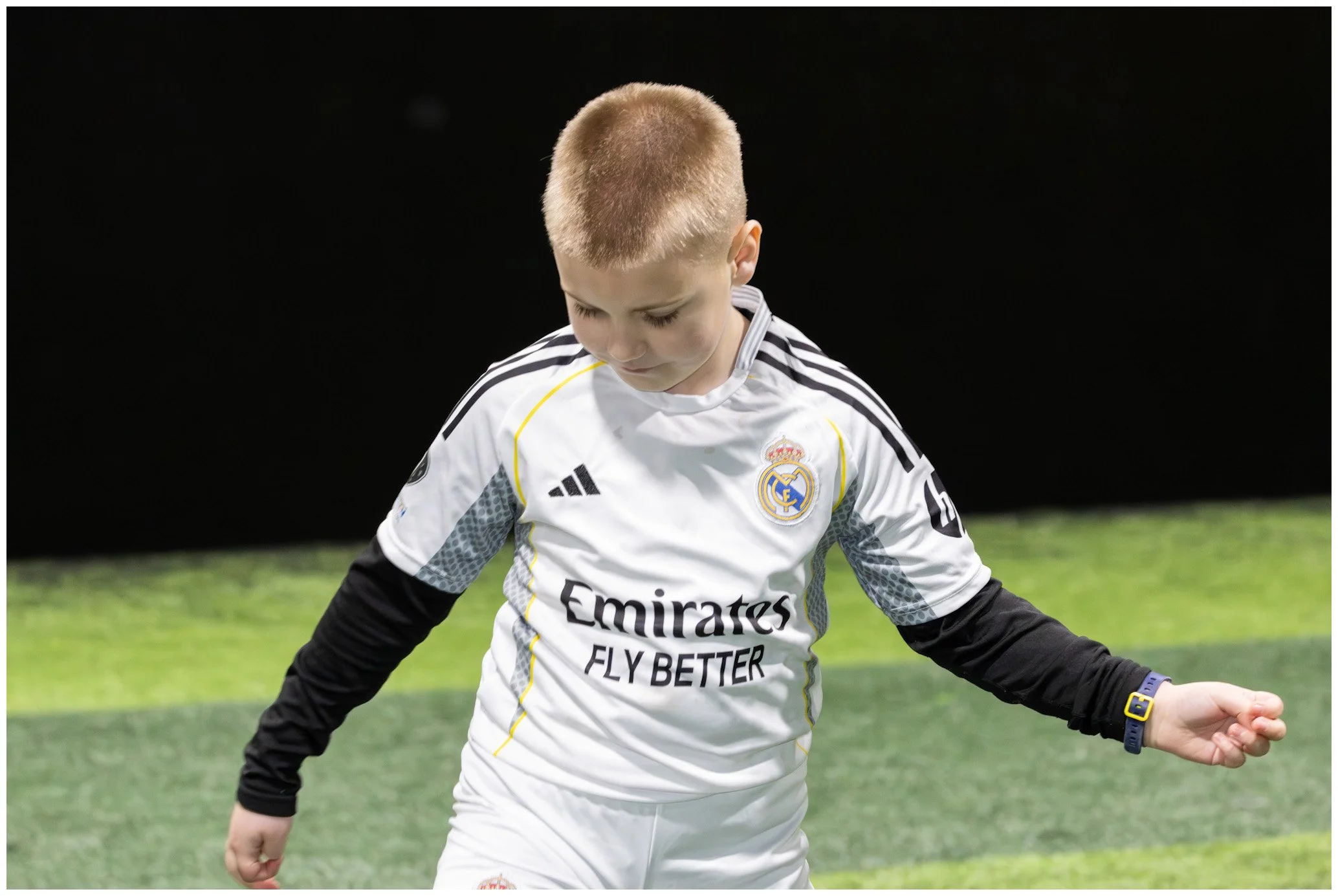 A young boy with short blonde hair wearing a Real Madrid soccer jersey standing on a field.