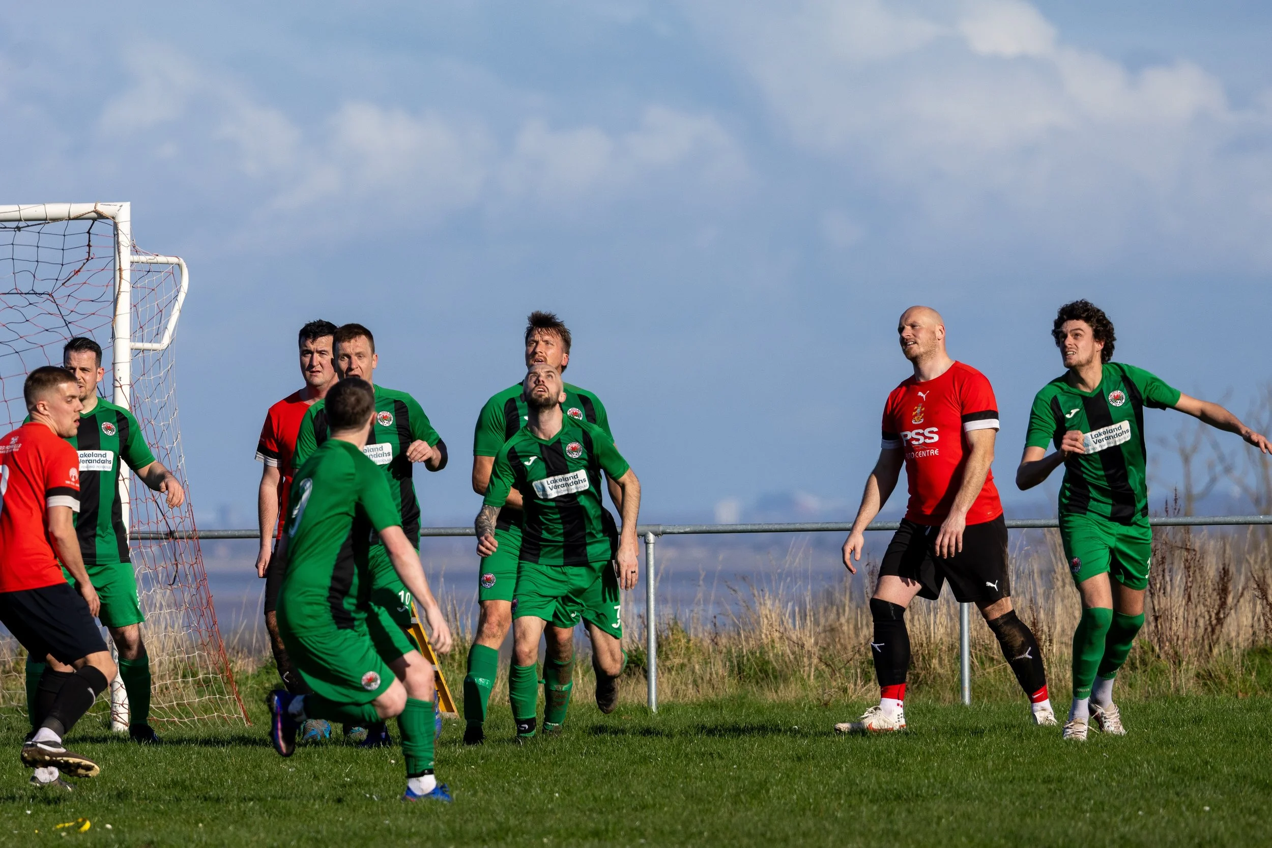 Soccer players in green and red jerseys on a grassy field near the goal, preparing for a shot during a game under a partly cloudy sky.