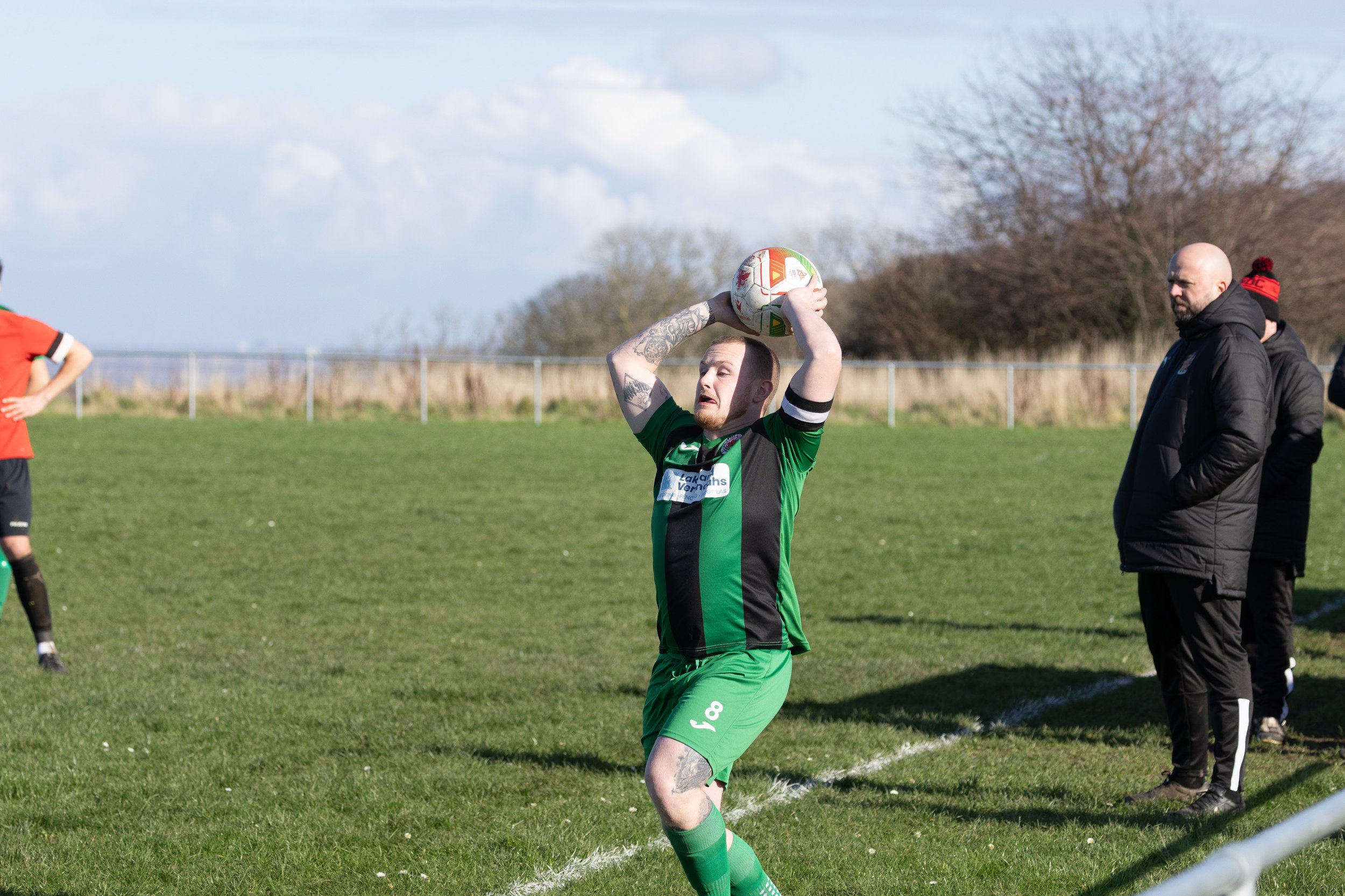 A rugby player in a green and black uniform preparing to throw the ball in a game, with a coach and another player in the background on a grassy field with a fence and trees in the distance.