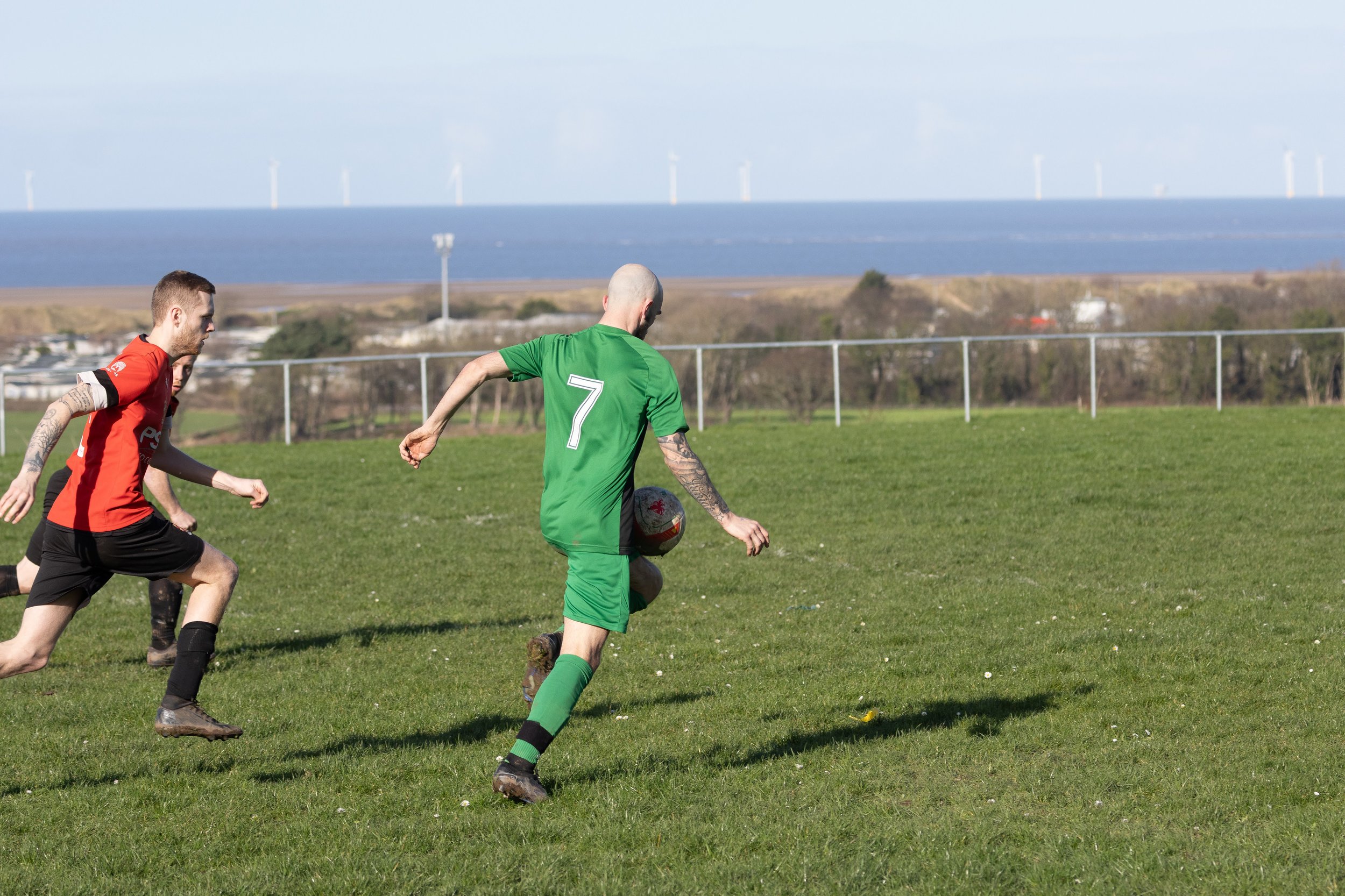 Two soccer players running on a grass field with a view of the ocean and wind turbines in the background.