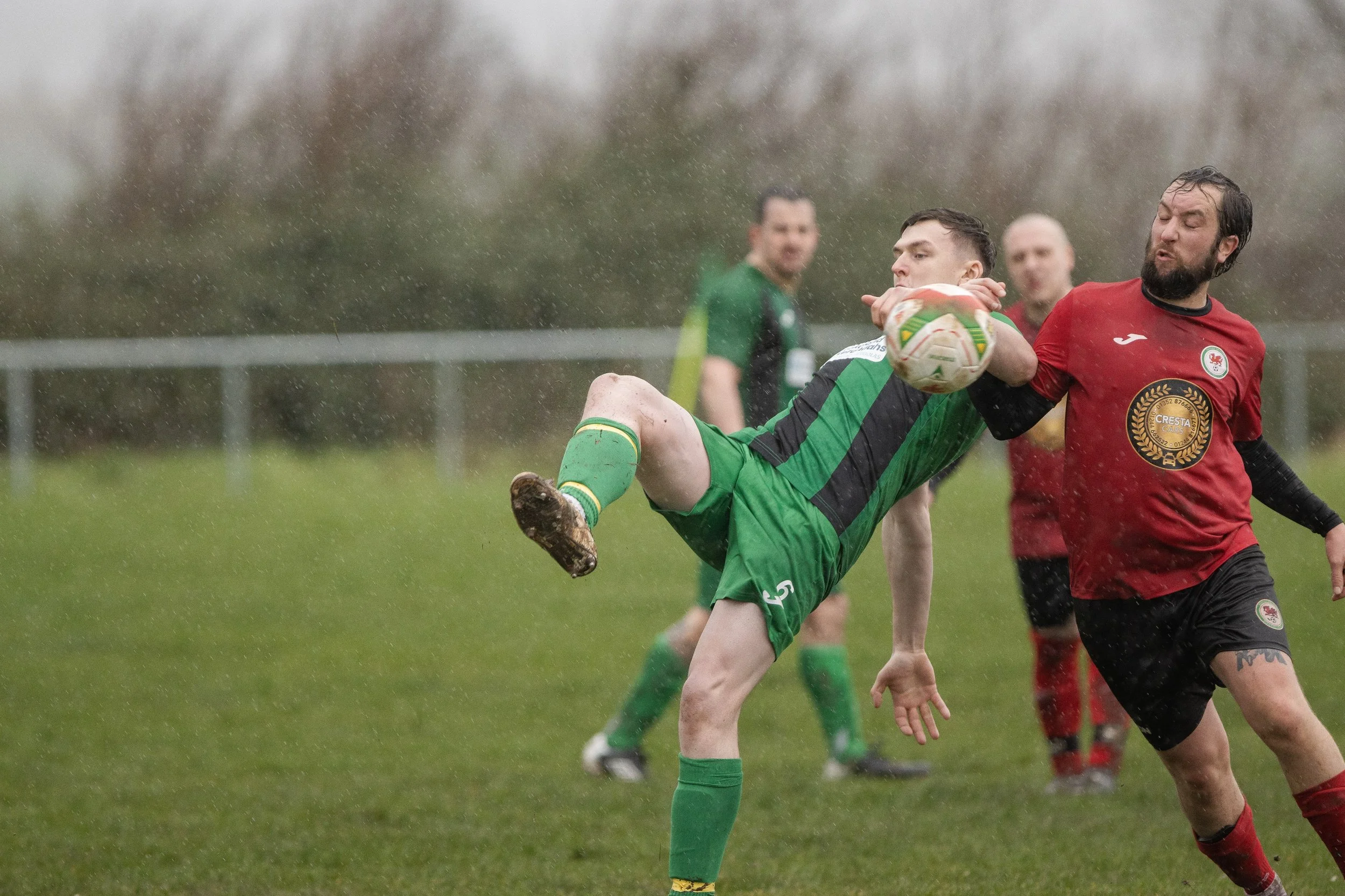 Two soccer players fighting for the ball in a rainy game, one wearing a green and black uniform and the other in a red uniform, with background players and overcast sky.